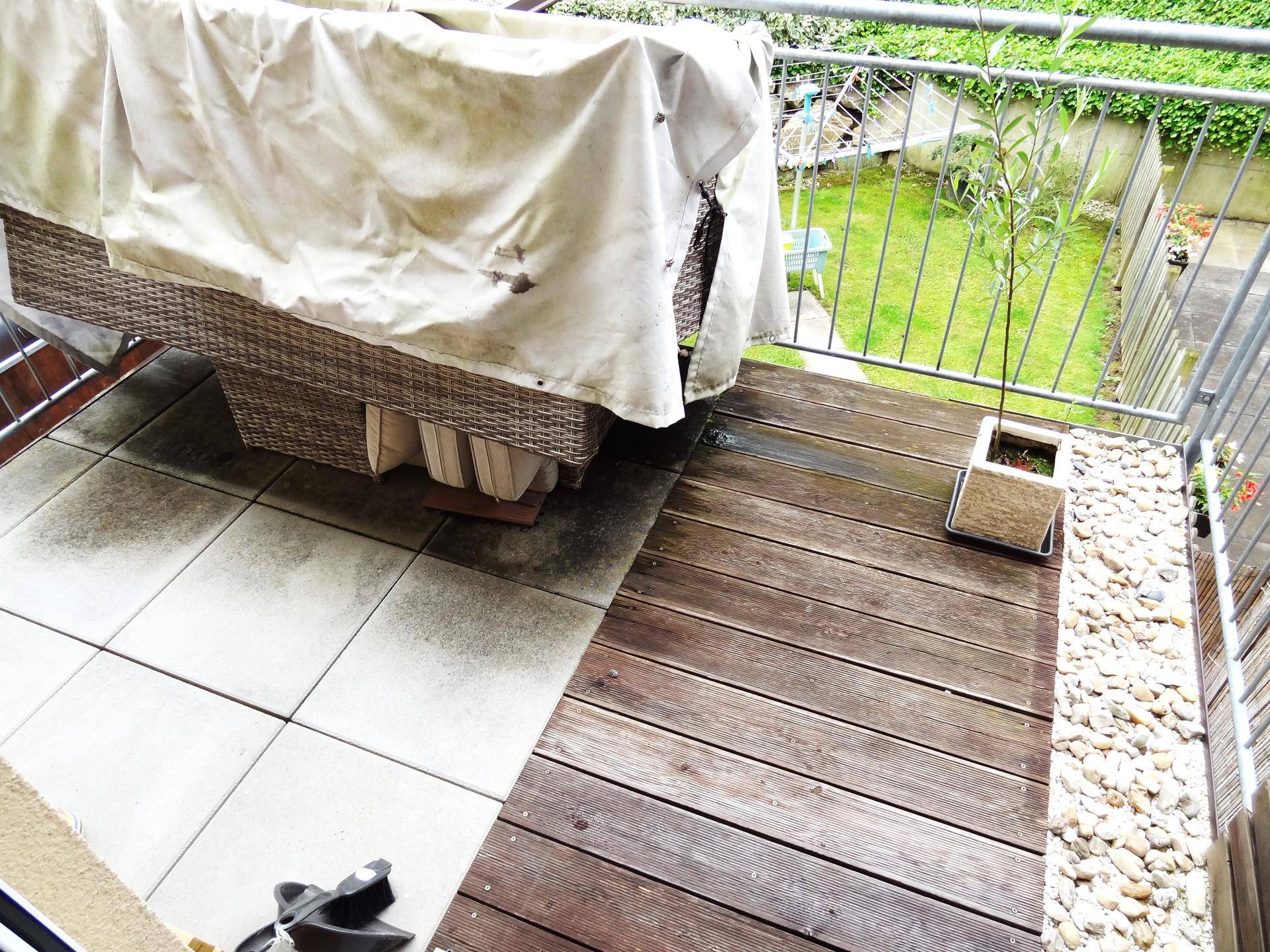 Geräumiger Balkon mit Holzdielen und Fliesen, Blick ins Grüne und abgedeckten Gartenmöbeln.