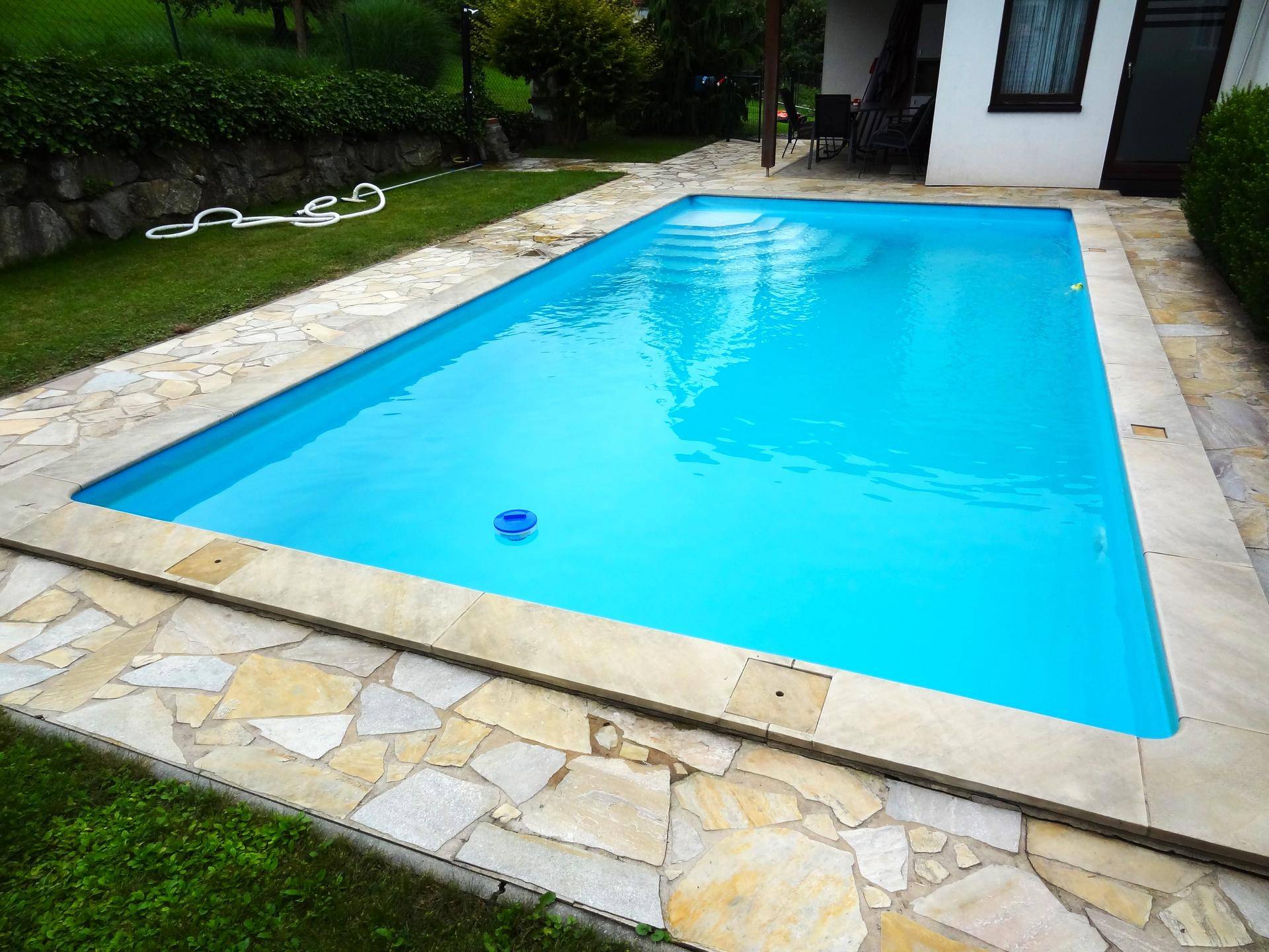 Rechteckiger Swimmingpool mit blauer Wasseroberfläche und Natursteinplatten im Garten.