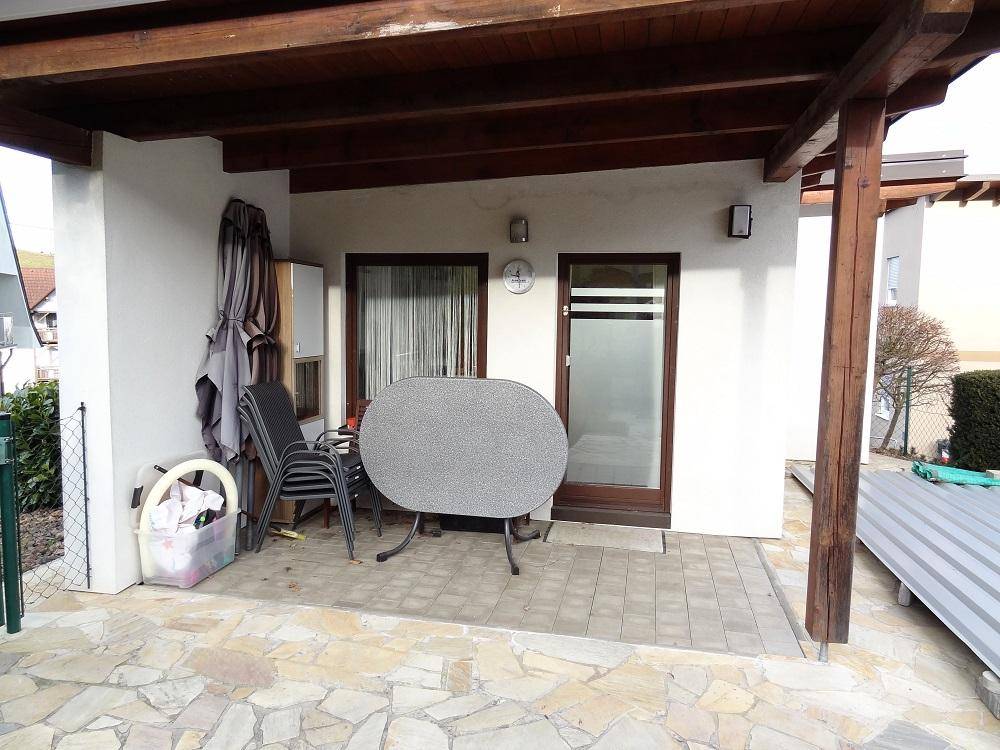 Überdachte Terrasse mit Holzbalken, Steinfliesen und einem Gartentisch für Mahlzeiten im Freien.