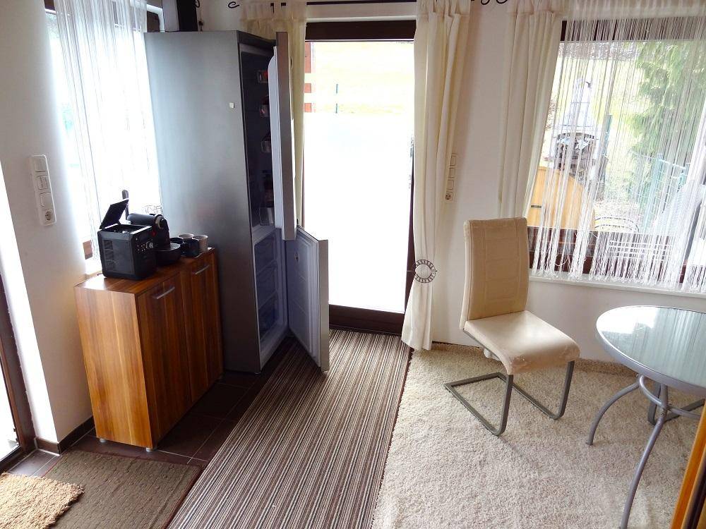 Bereich mit Kühlschrank und Sitzgelegenheiten, hell durch große Fenster und Glastür.