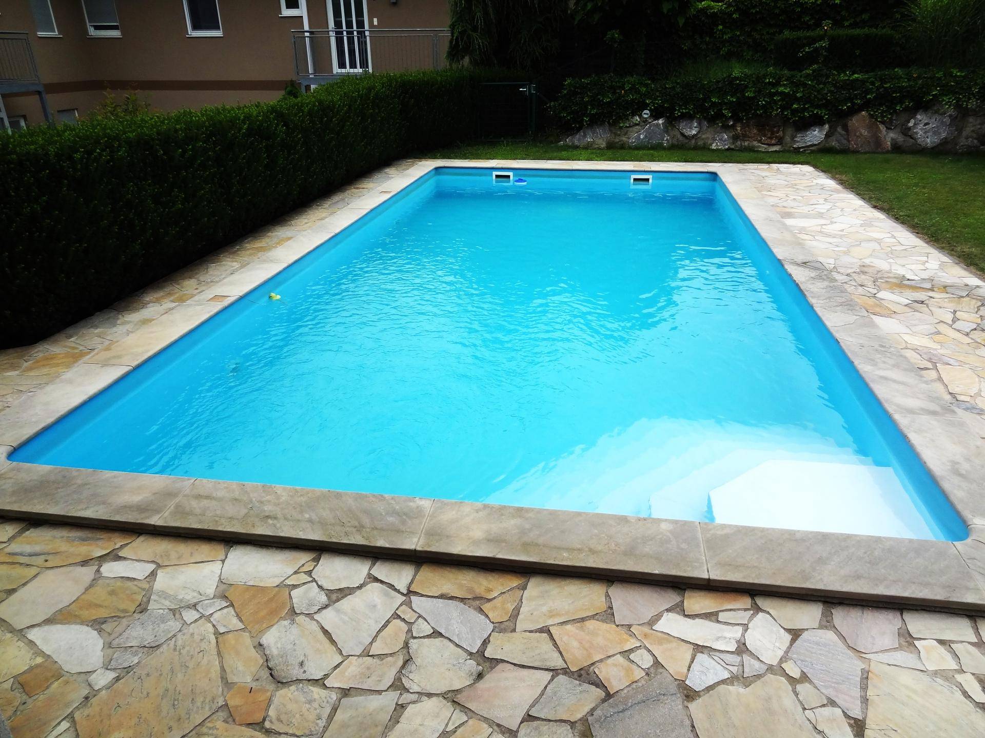 Blauer Swimmingpool mit integrierten Stufen und breitem Rand aus Natursteinplatten.