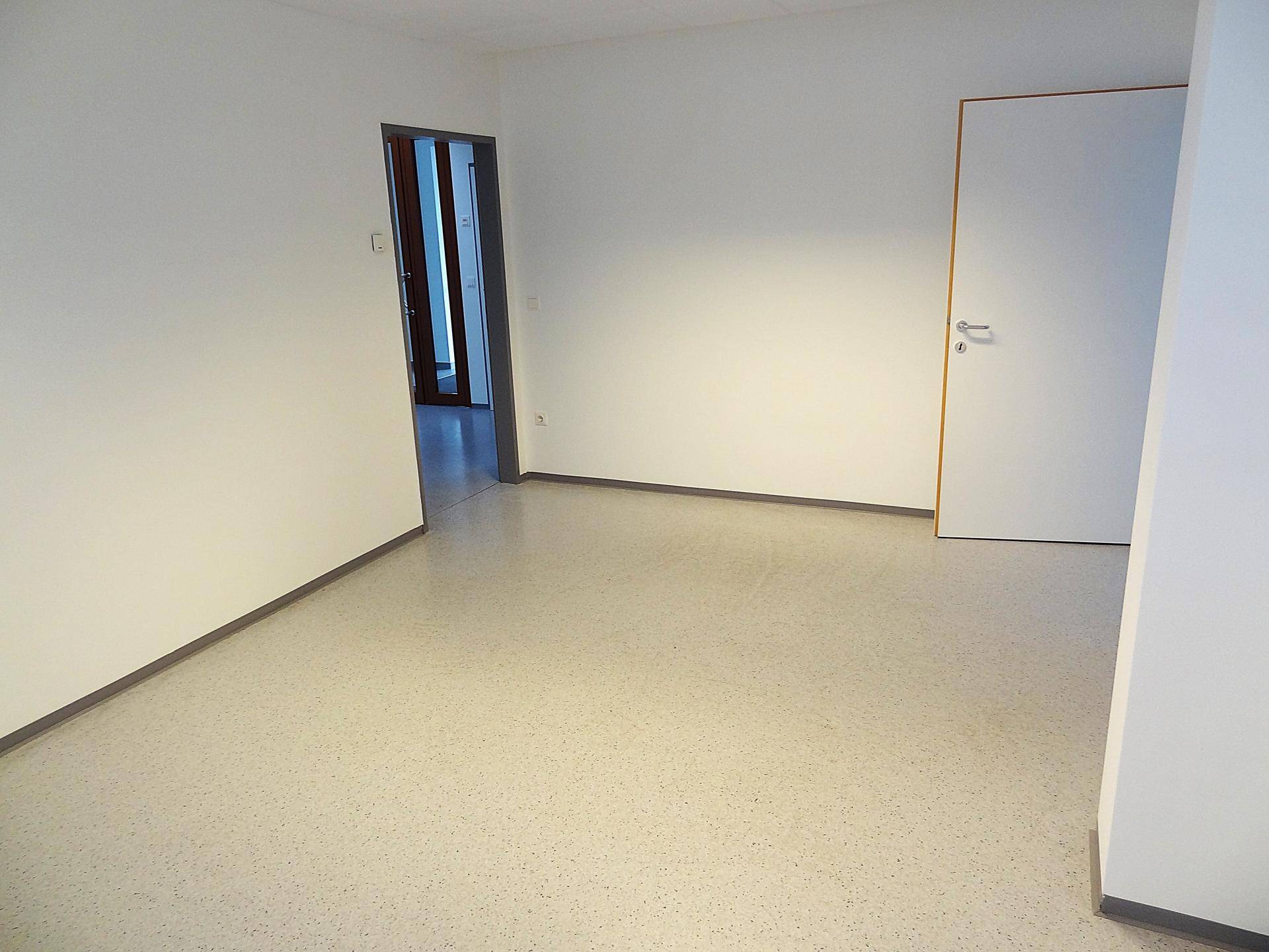 Unmöblierter Raum mit hellem Vinylboden und einer offenen Tür, die den Blick in einen weiteren Flur freigibt.