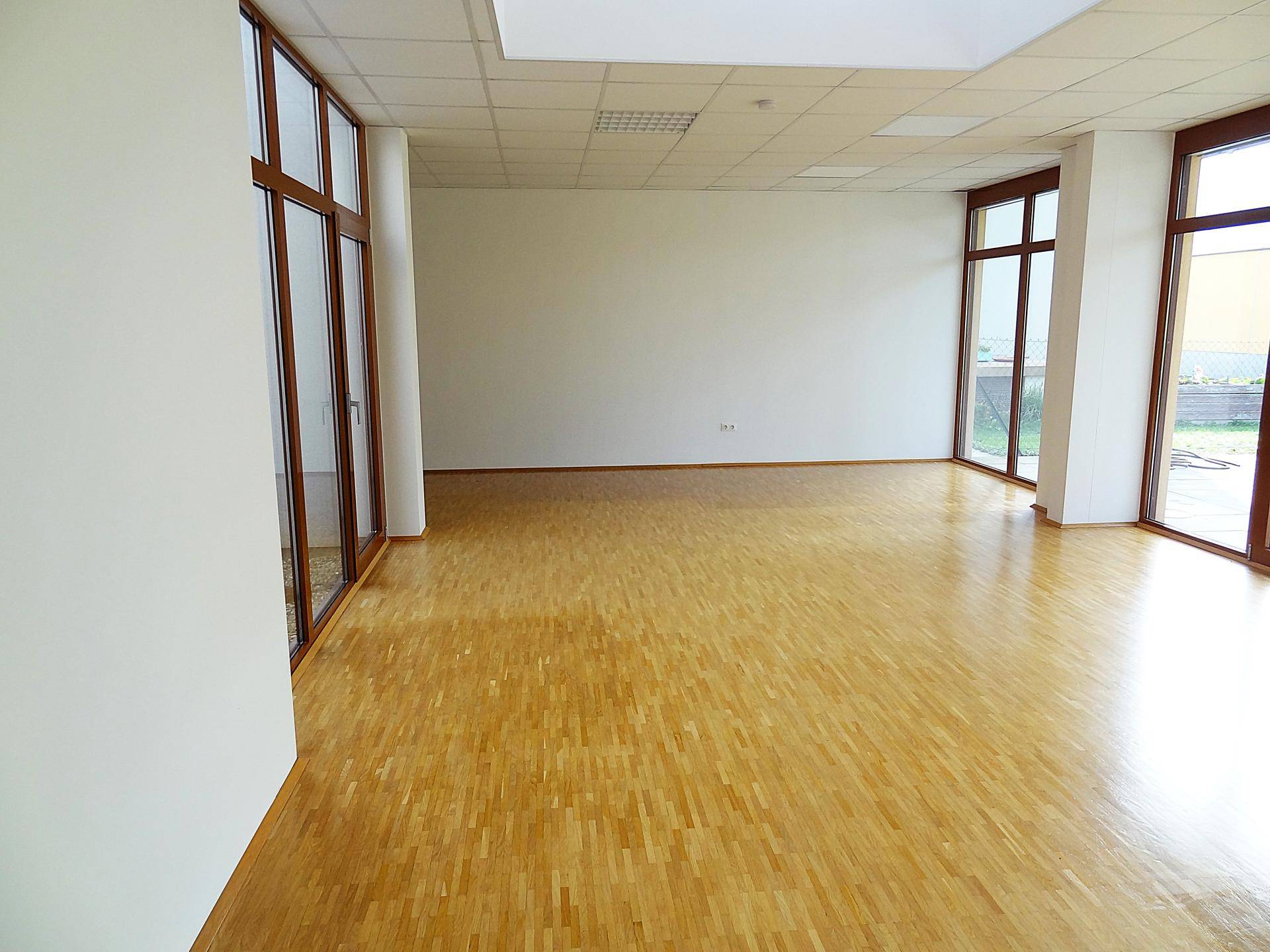 Geräumiger, unmöblierter Raum mit glänzendem Parkettboden und großen Fenstern, die den Blick nach draußen freigeben.