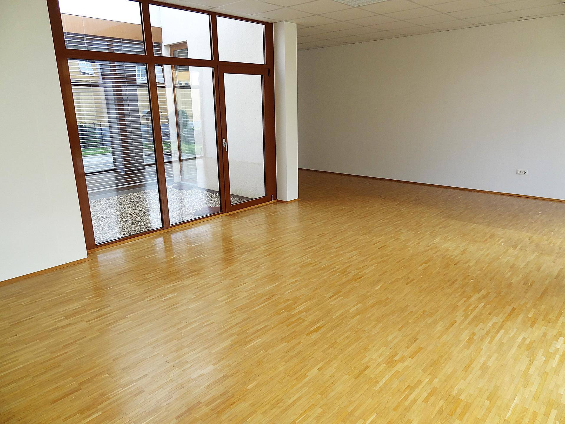 Großer, heller Raum mit Parkettboden und breiten Fenstern, die einen Blick auf einen Innenhof ermöglichen.