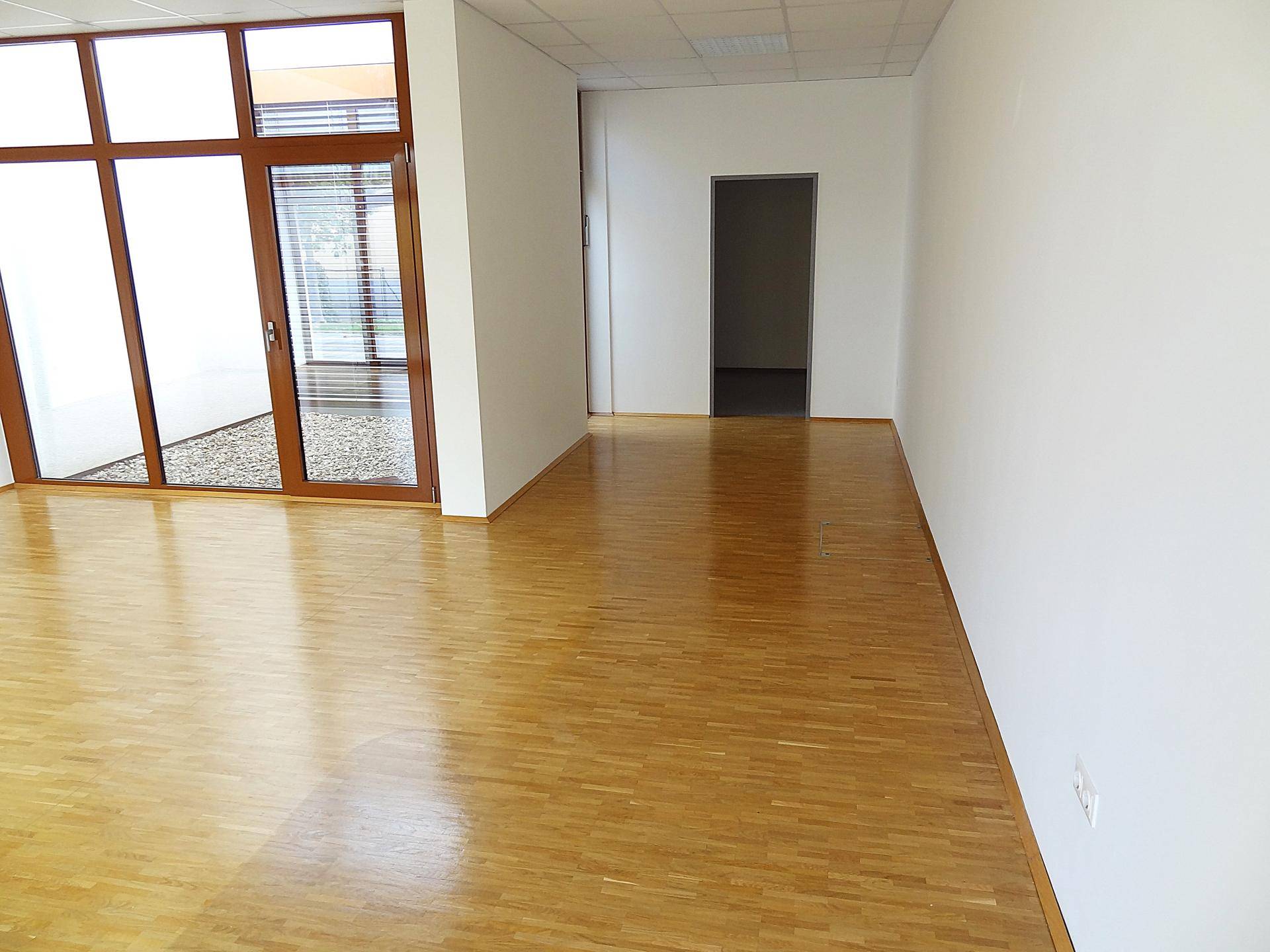 Heller Raum mit Parkettboden und einer Tür, die in einen weiteren Bereich führt, sowie großen Fenstern.