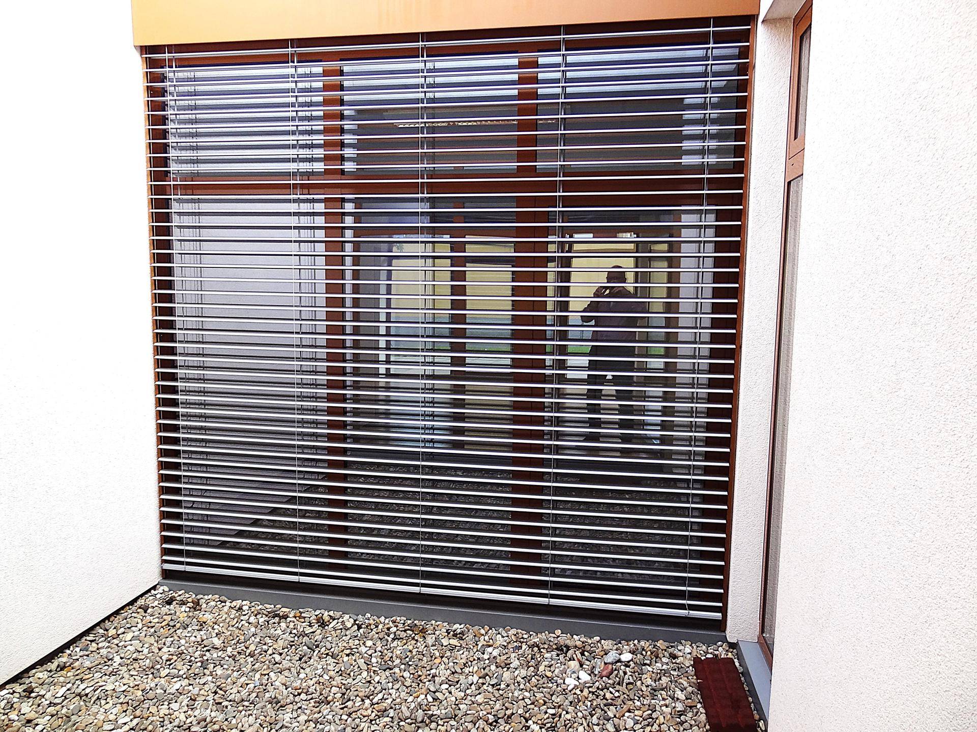 Außenansicht eines großen Fensters mit Jalousien und einem Kiesbeet davor.