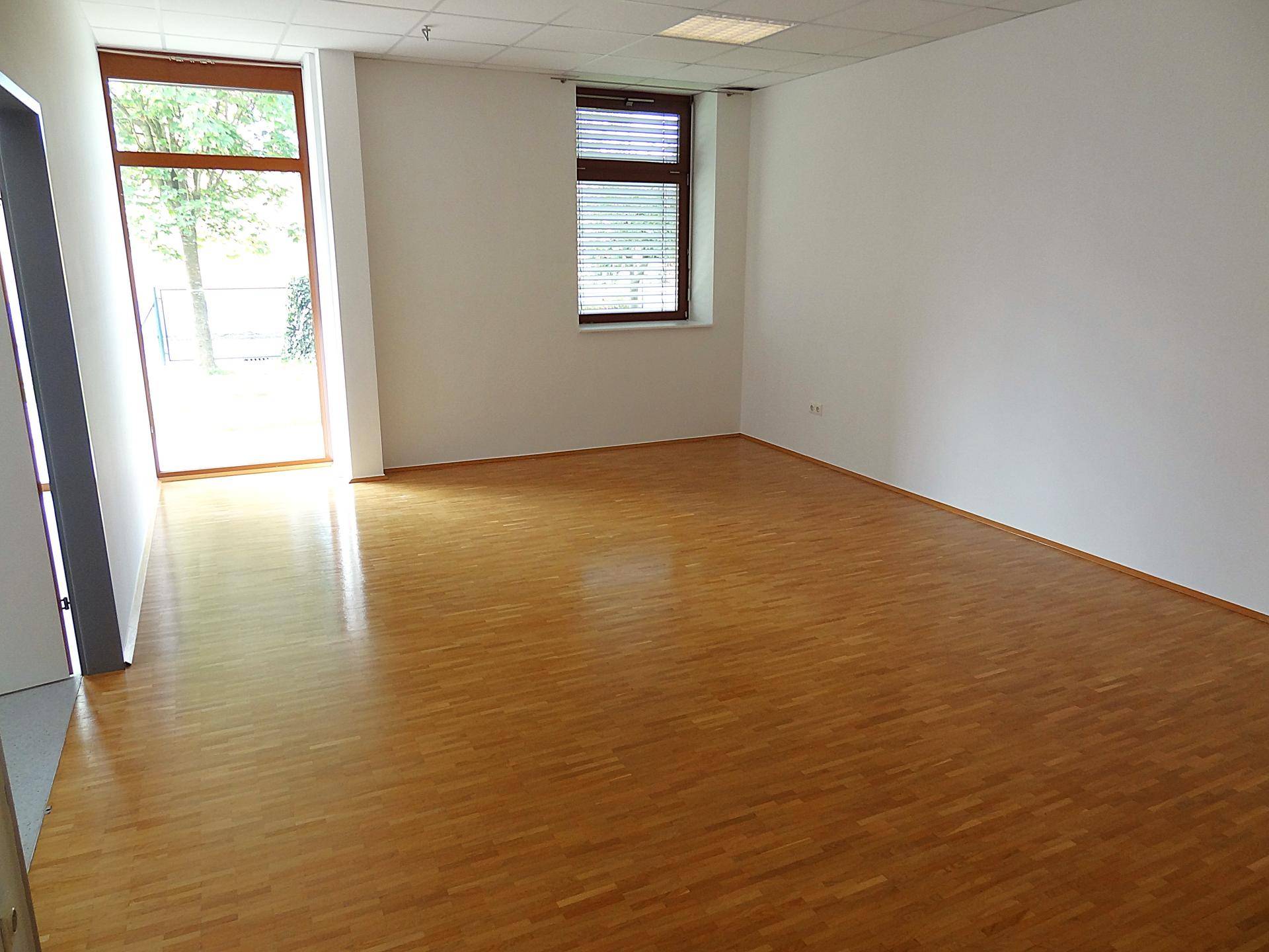 Geräumiger Büroraum mit Holzboden, Fenster mit Jalousien und viel Tageslicht.