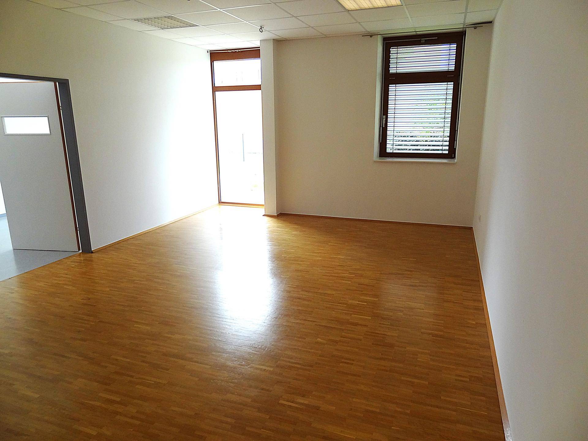 Heller Büroraum mit Holzboden, Fenster mit Jalousien und Zugang zu einem angrenzenden Bereich.