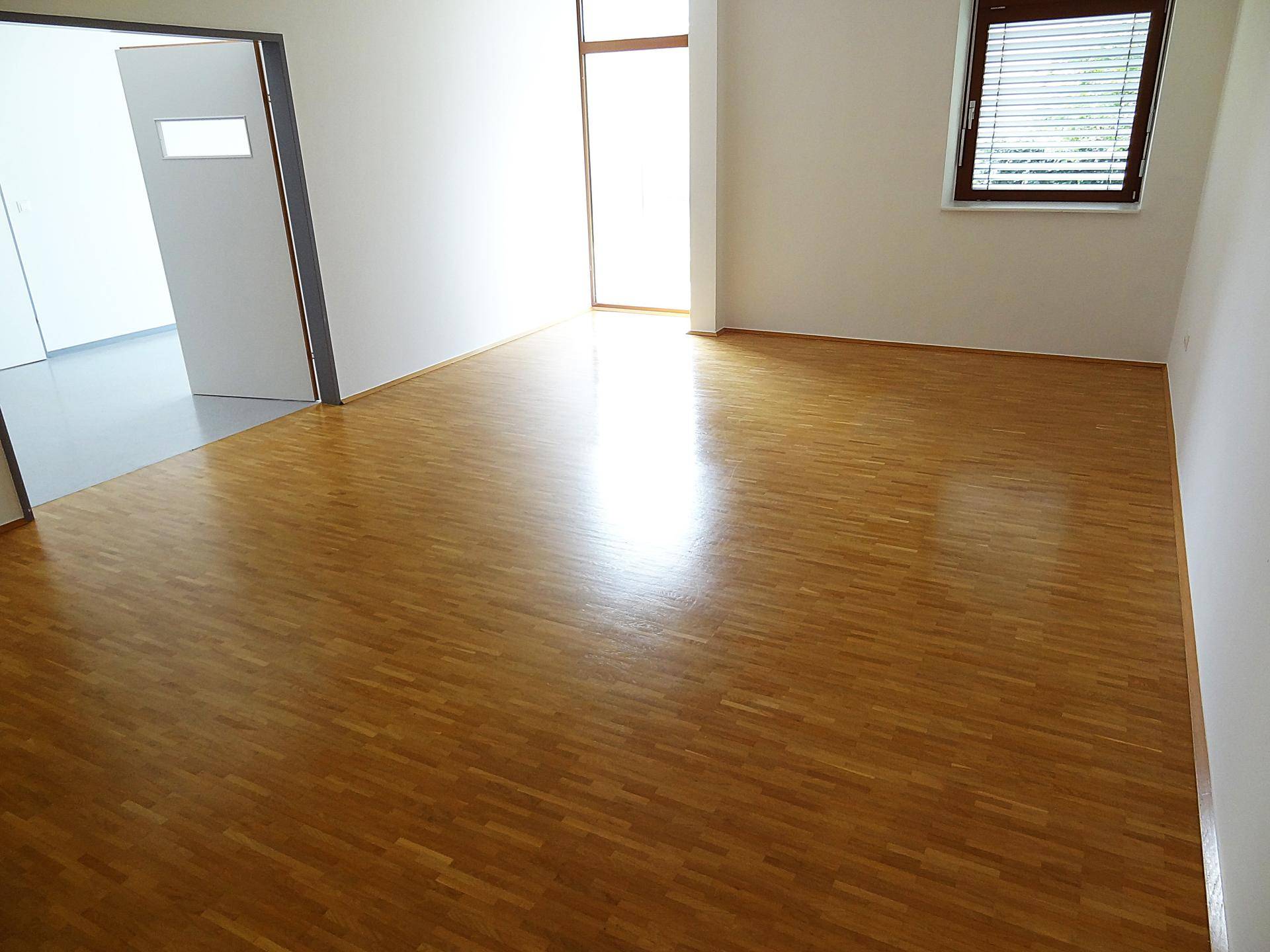 Sonniger Büroraum mit glänzendem Holzboden und Fenster mit Jalousien.