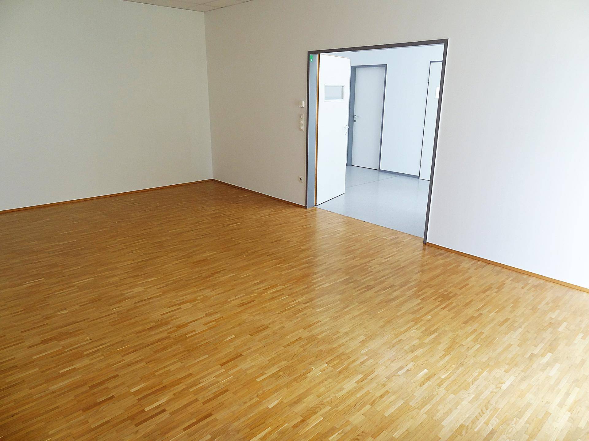 Leerer Büroraum mit Holzboden und offener Tür zu einem angrenzenden Bereich.