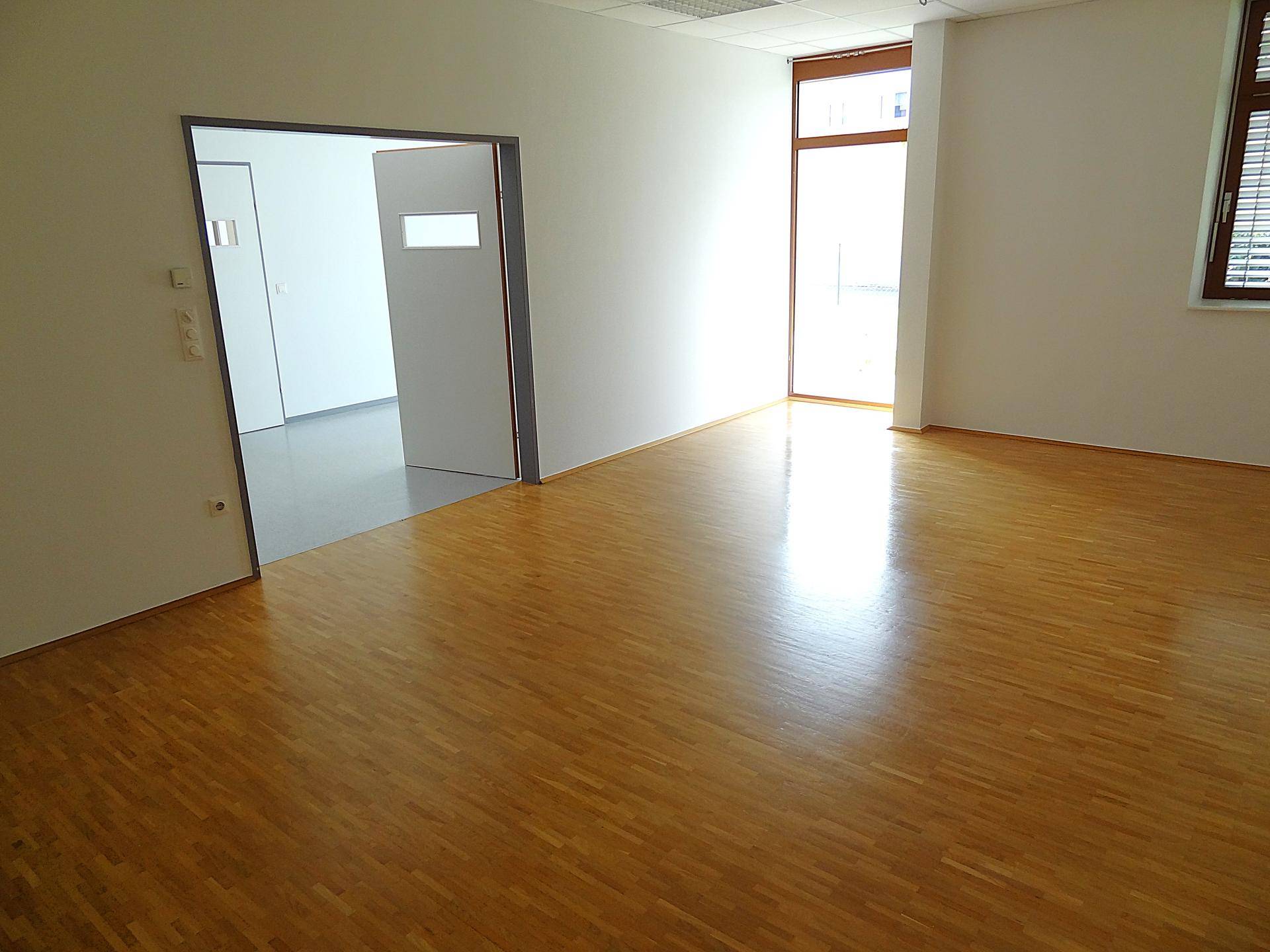 Heller Büroraum mit Holzboden, Fenster mit Jalousien und offener Tür.