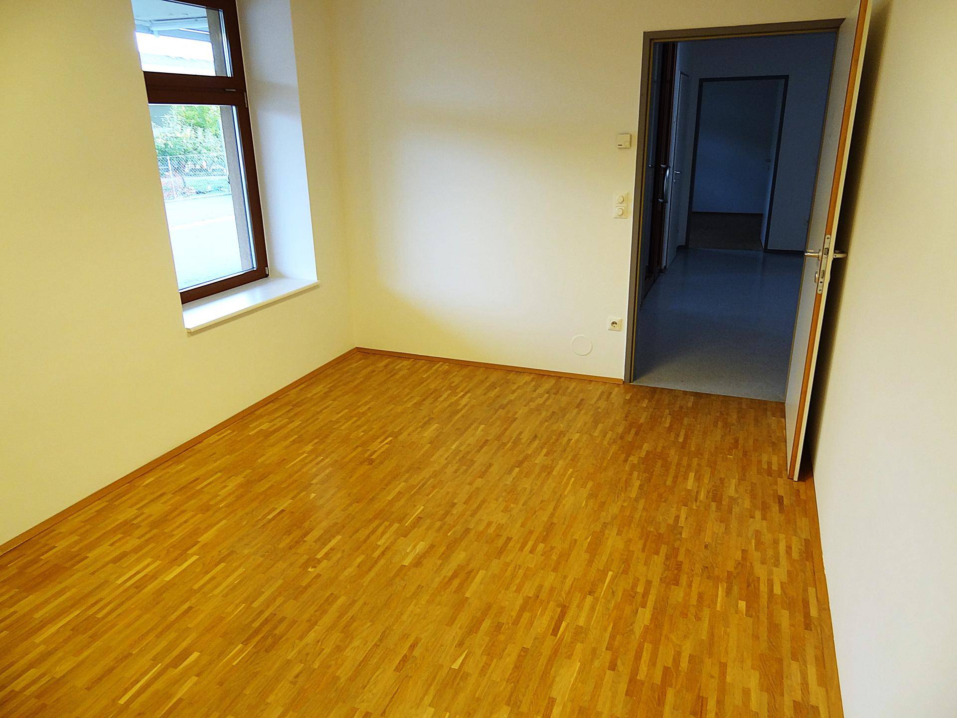 Heller Büroraum mit Holzboden, Fenster und offener Tür zu einem Flur.