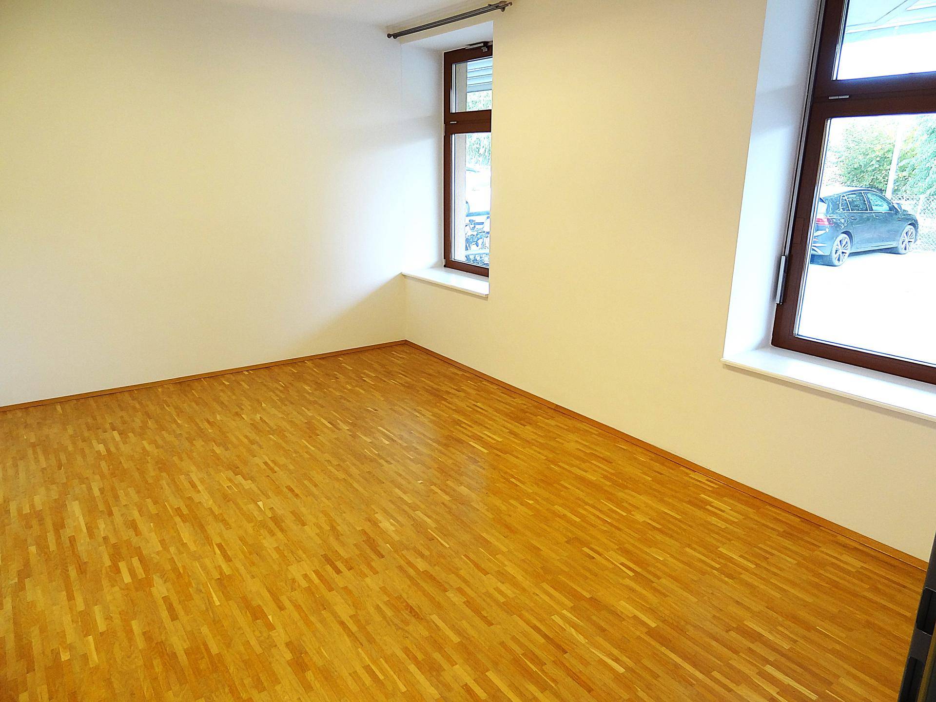 Leerer Büroraum mit Holzboden und zwei Fenstern, eines davon mit Jalousien.