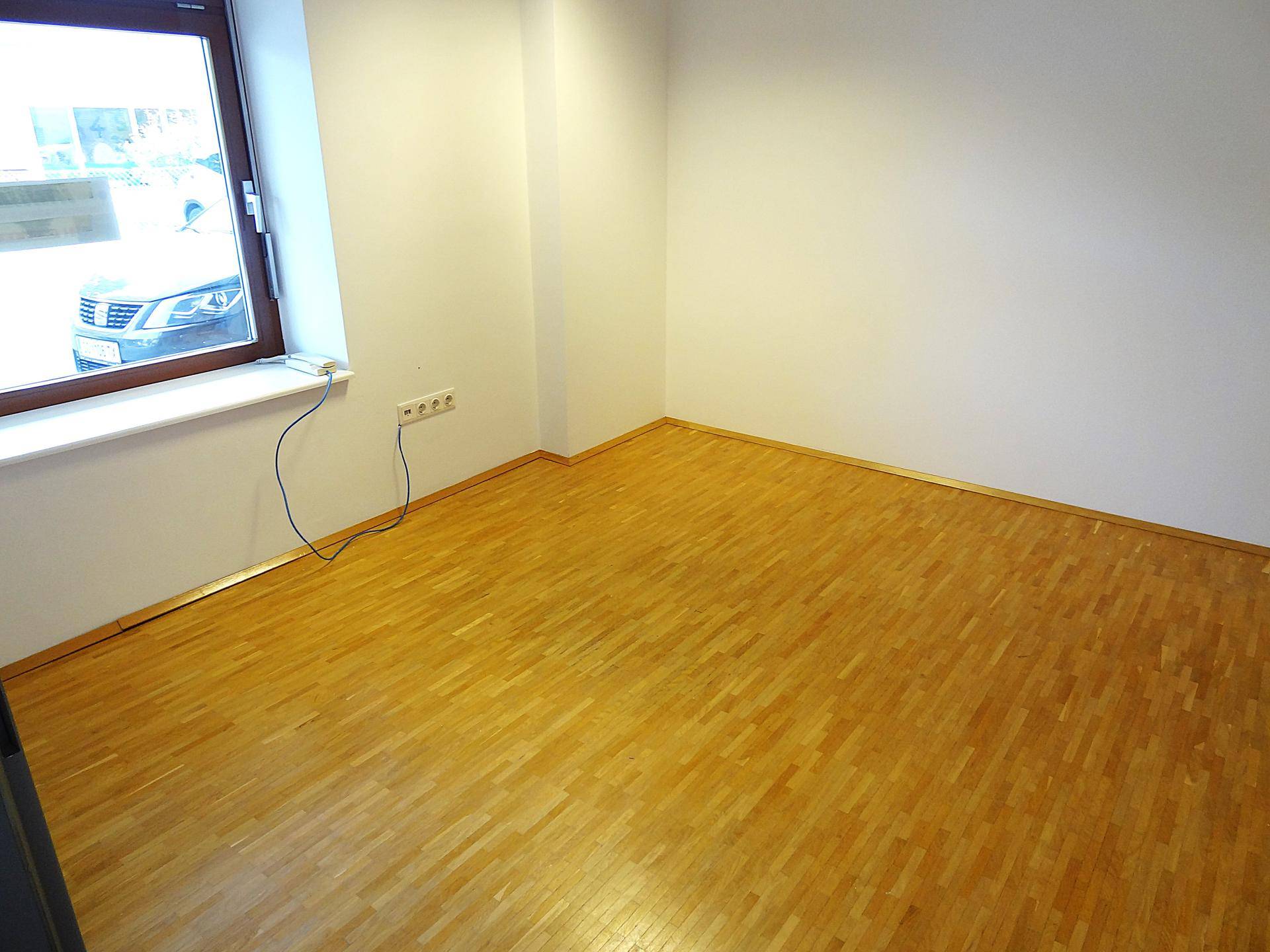 Büroraum mit Holzboden, Fenster und sichtbaren Steckdosen an der Wand.