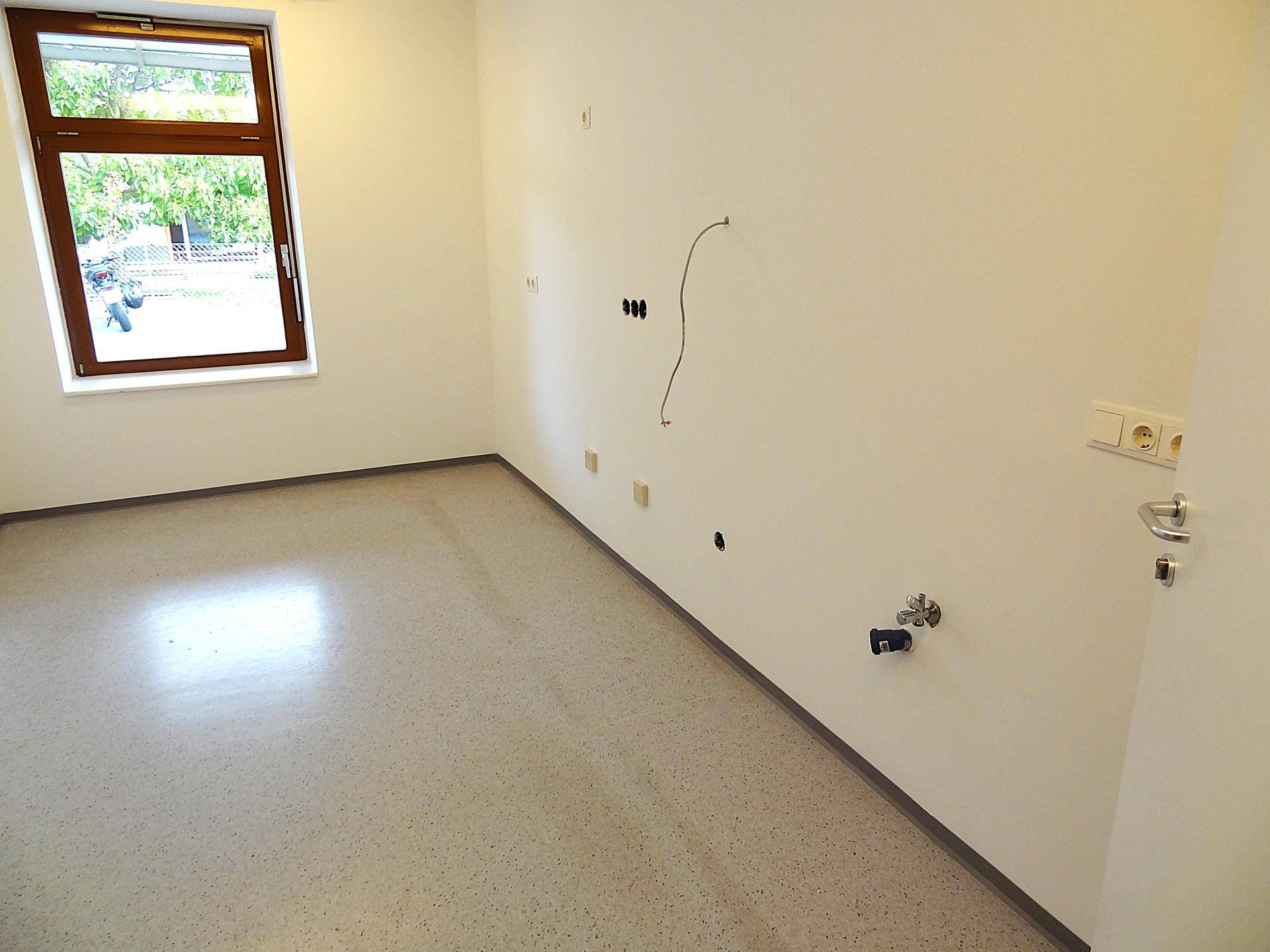 Raum mit hellem Boden, Fenster und vorbereiteten Anschlüssen an der Wand.