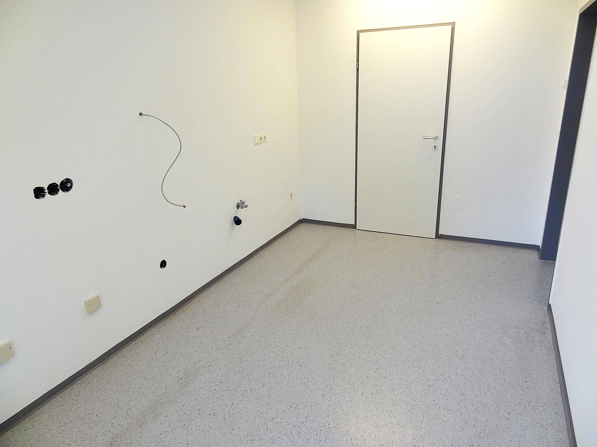 Raum mit hellem Boden, vorbereiteten Anschlüssen an der Wand und einer Tür.