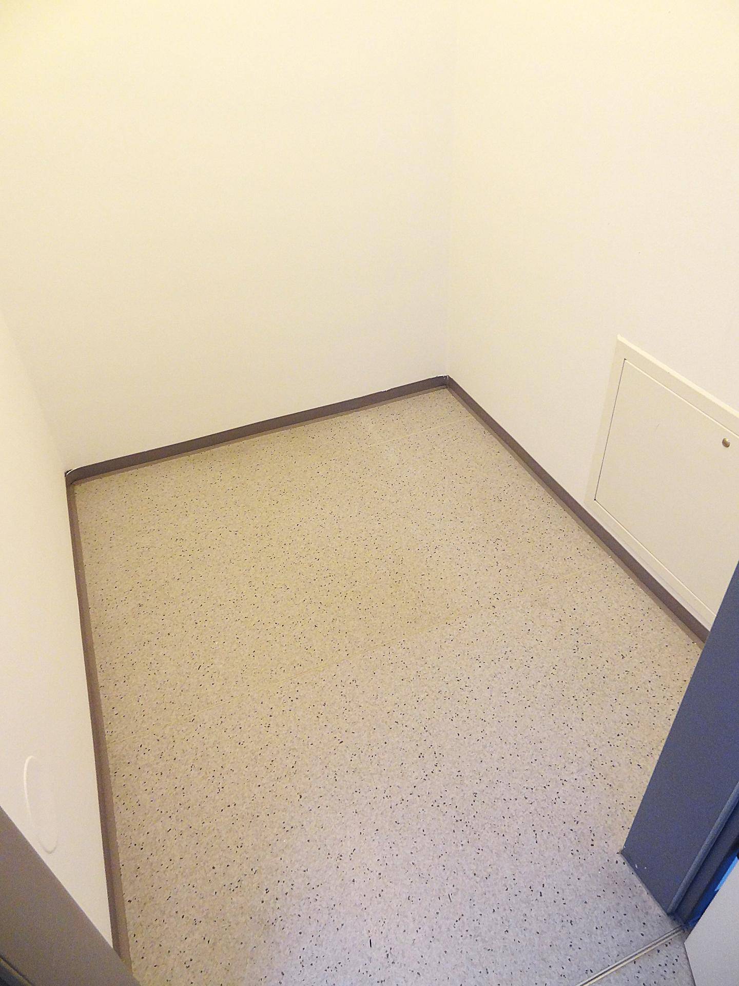 Kleiner, leerer Raum mit hellem Boden und einer Wartungsklappe an der Wand.