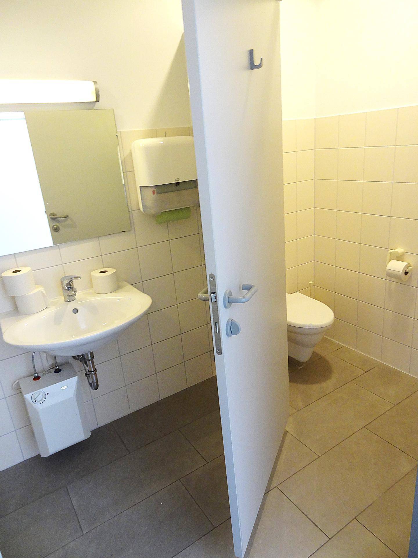 Badezimmer mit Waschbecken, Spiegel und offener Tür zur Toilette, hell gefliest.
