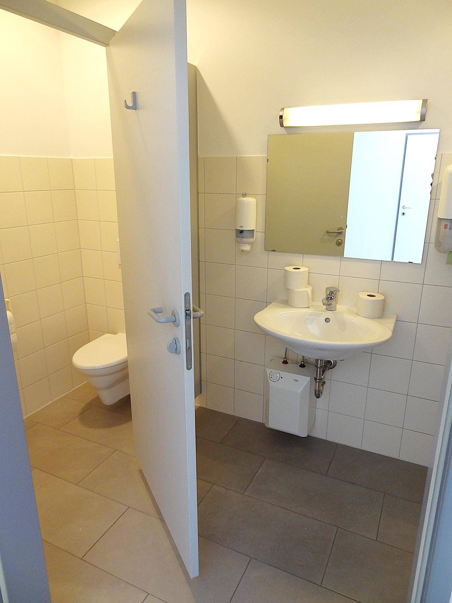Badezimmer mit Waschbecken, Spiegel und offener Tür zur Toilette, hell gefliest.
