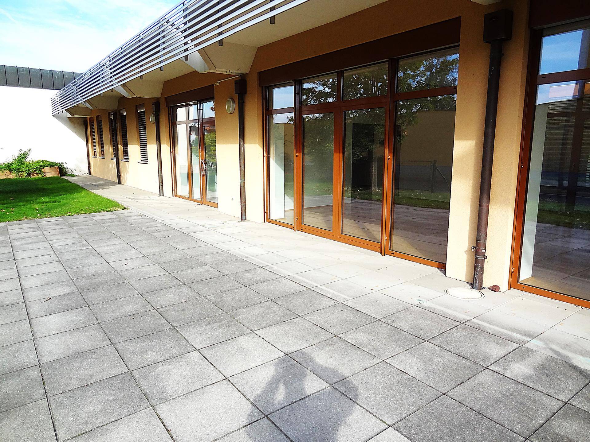 Weitläufige gepflasterte Terrasse entlang der Gebäudefassade mit mehreren großen Fenstern.