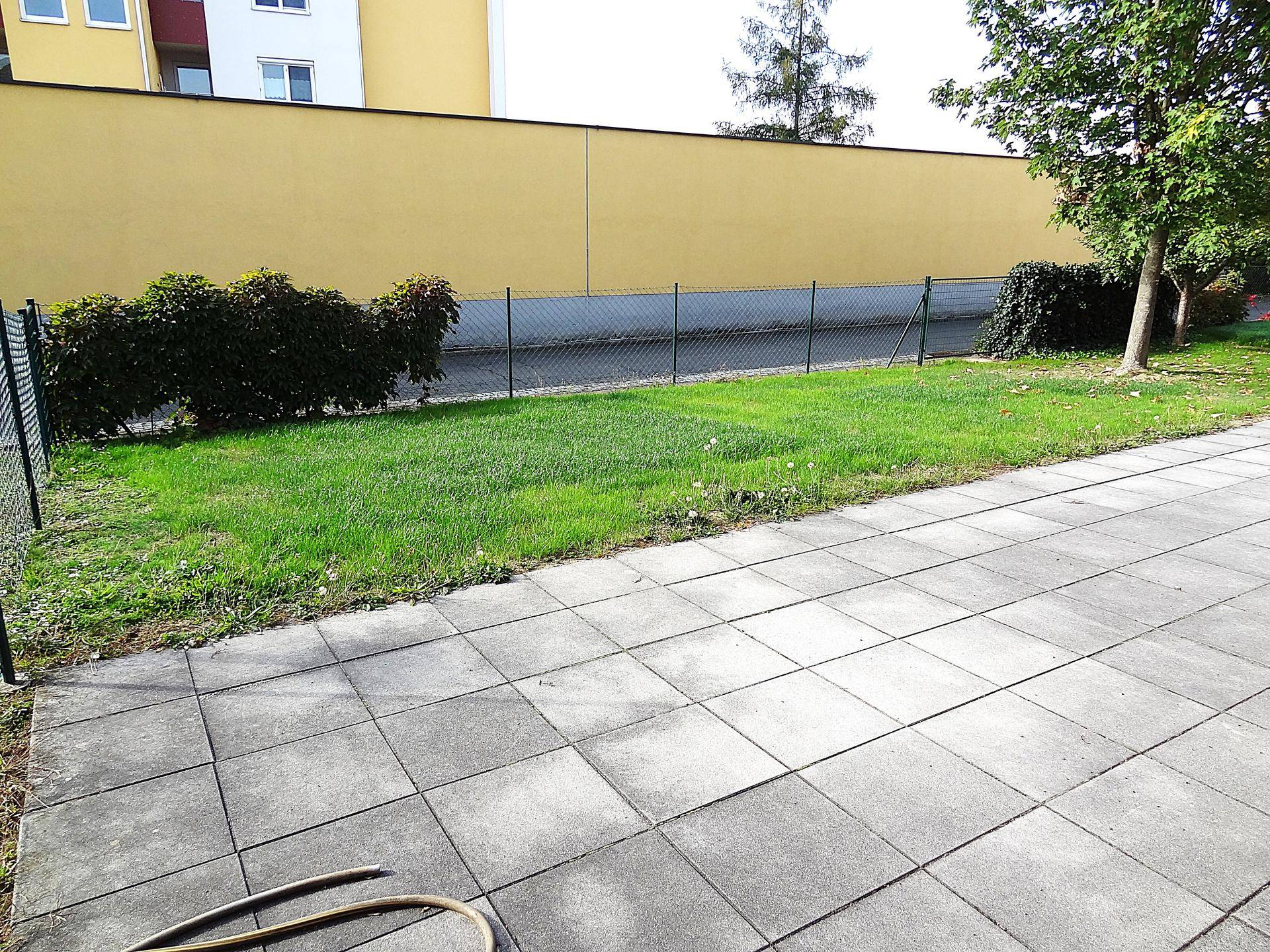 Gepflegte Rasenfläche mit Zaun und angrenzender gepflasterter Terrasse vor einem Gebäude.