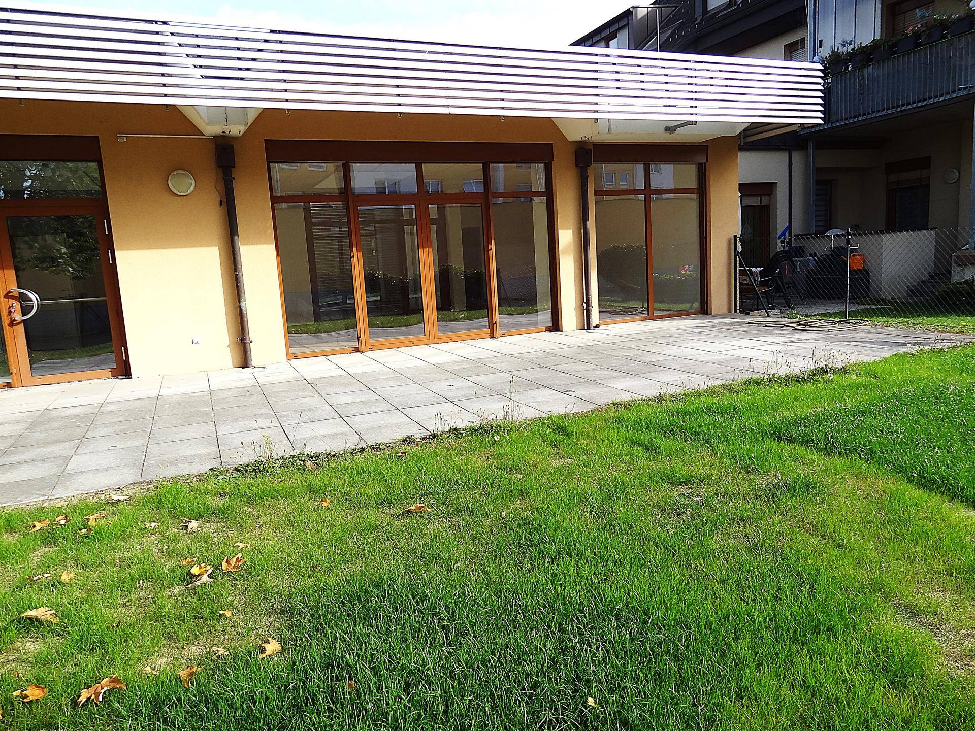 Gebäudefassade mit großen Fenstern und einer gepflasterten Terrasse, umgeben von Rasen.