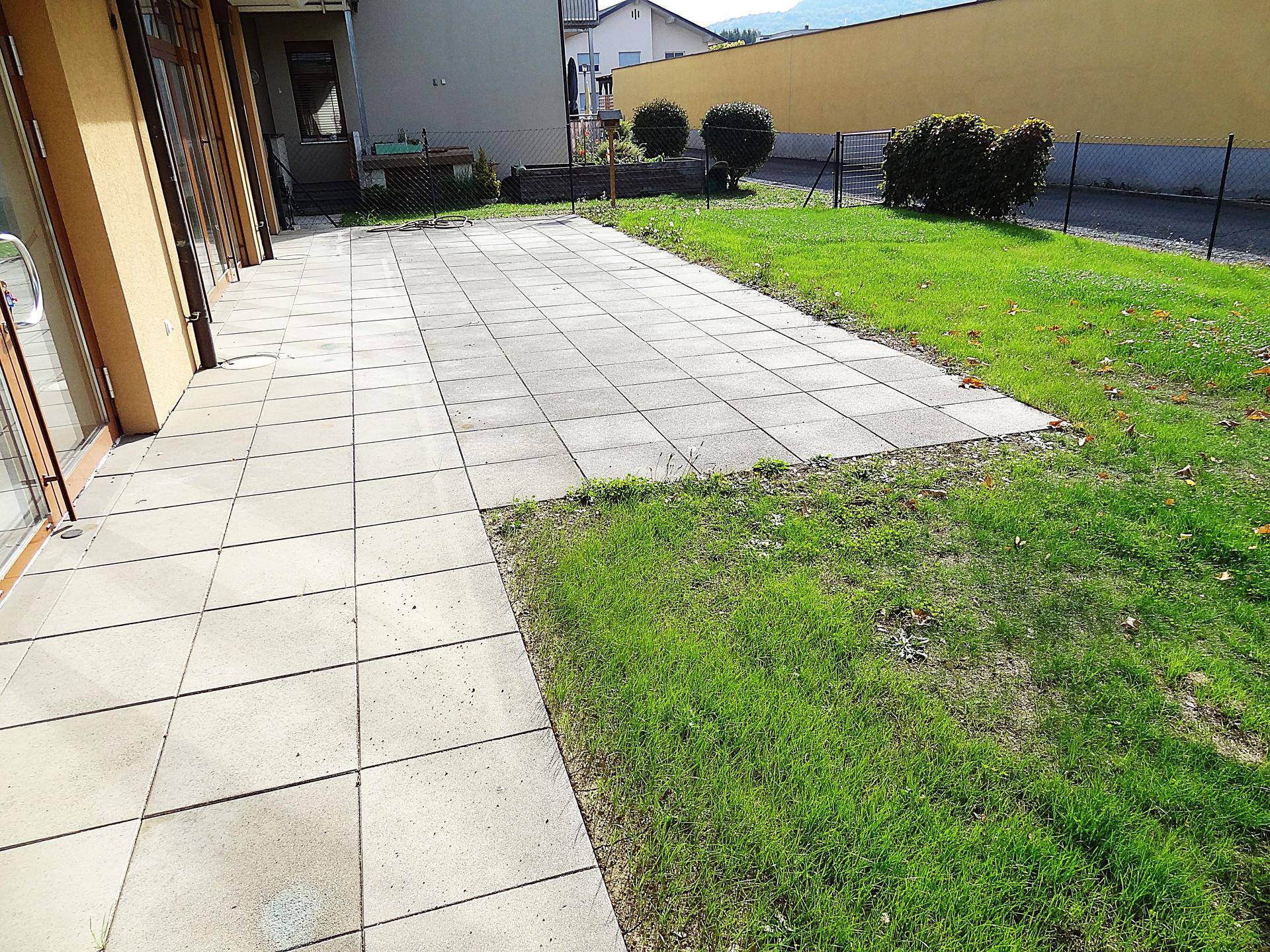 Weitläufige gepflasterte Terrasse neben einer Rasenfläche mit Blick auf angrenzende Gebäude.