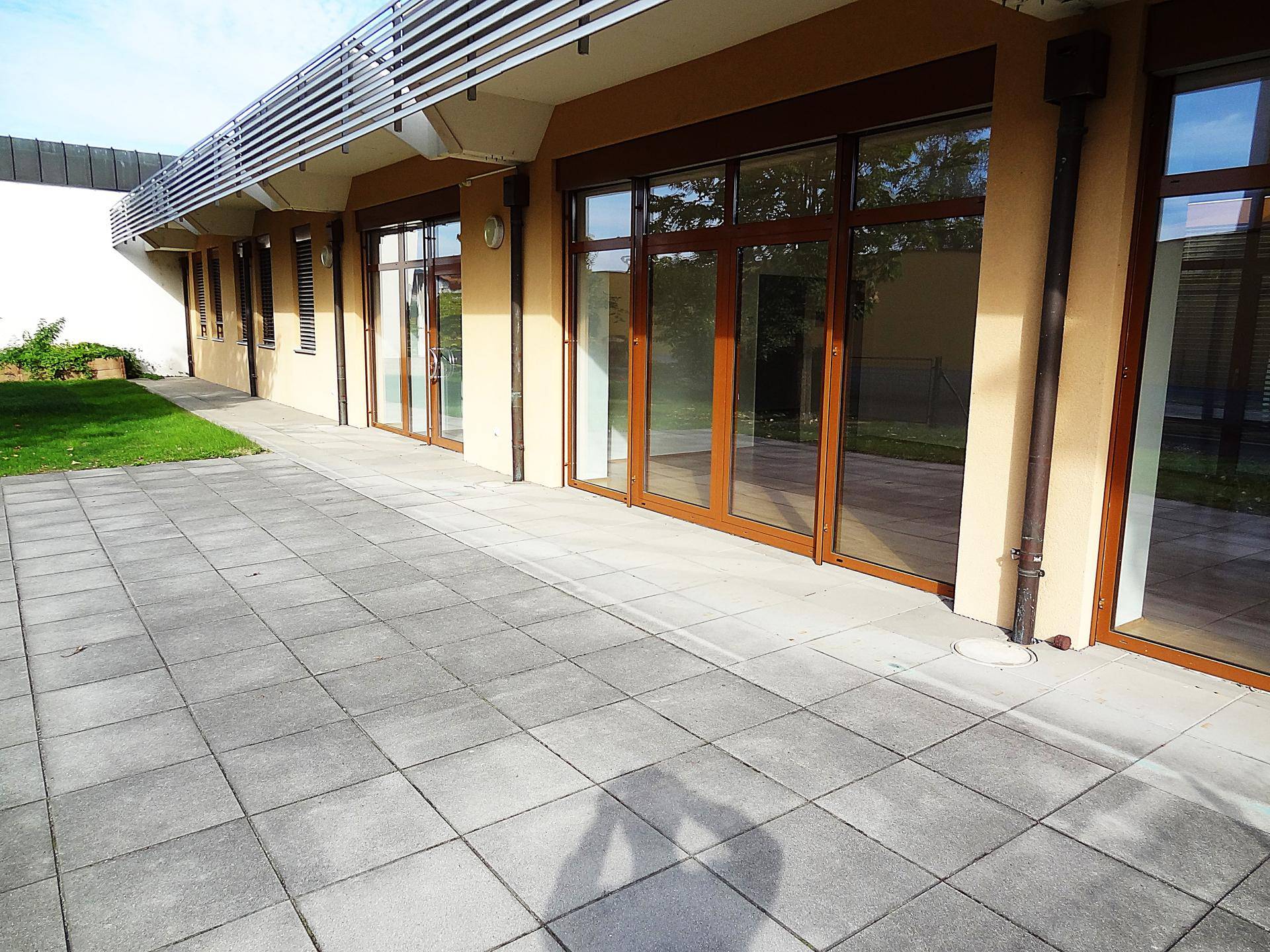 Weitläufige gepflasterte Terrasse entlang der Gebäudefassade mit mehreren großen Fenstern.