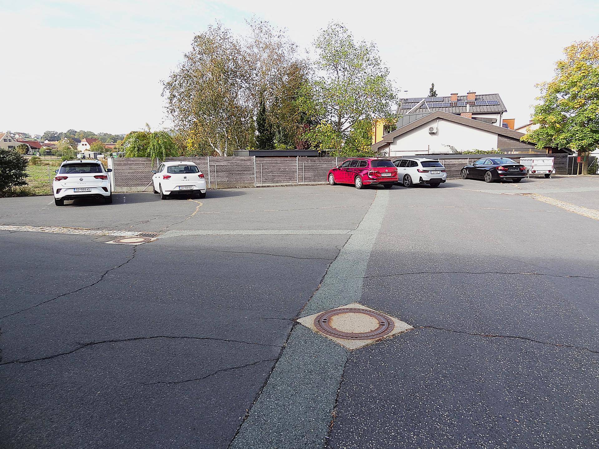 Weitläufiger asphaltierter Parkplatz mit geparkten Fahrzeugen und Gebäuden im Hintergrund.