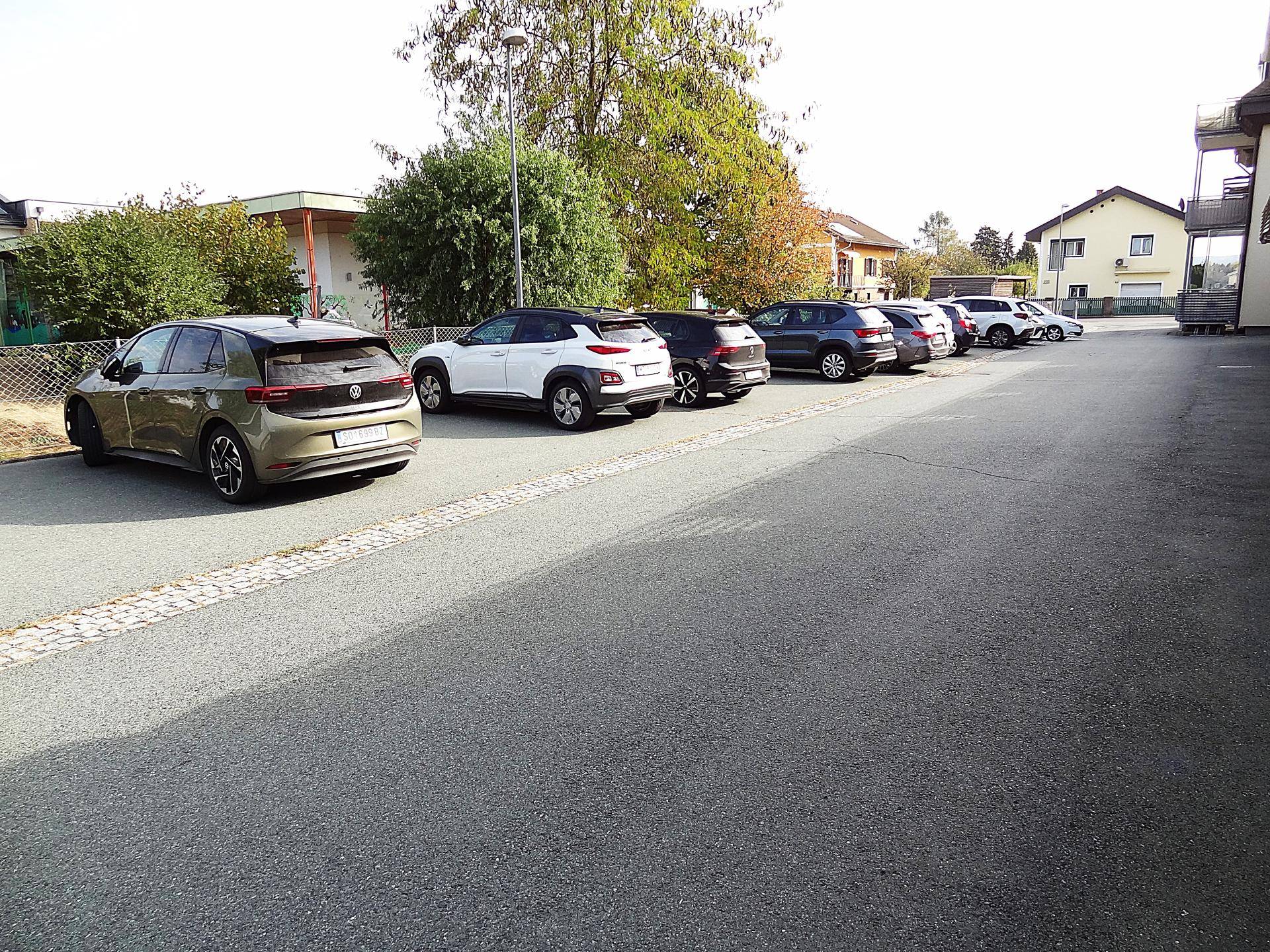 Asphaltierter Parkplatz mit mehreren geparkten Fahrzeugen und Bäumen im Hintergrund.