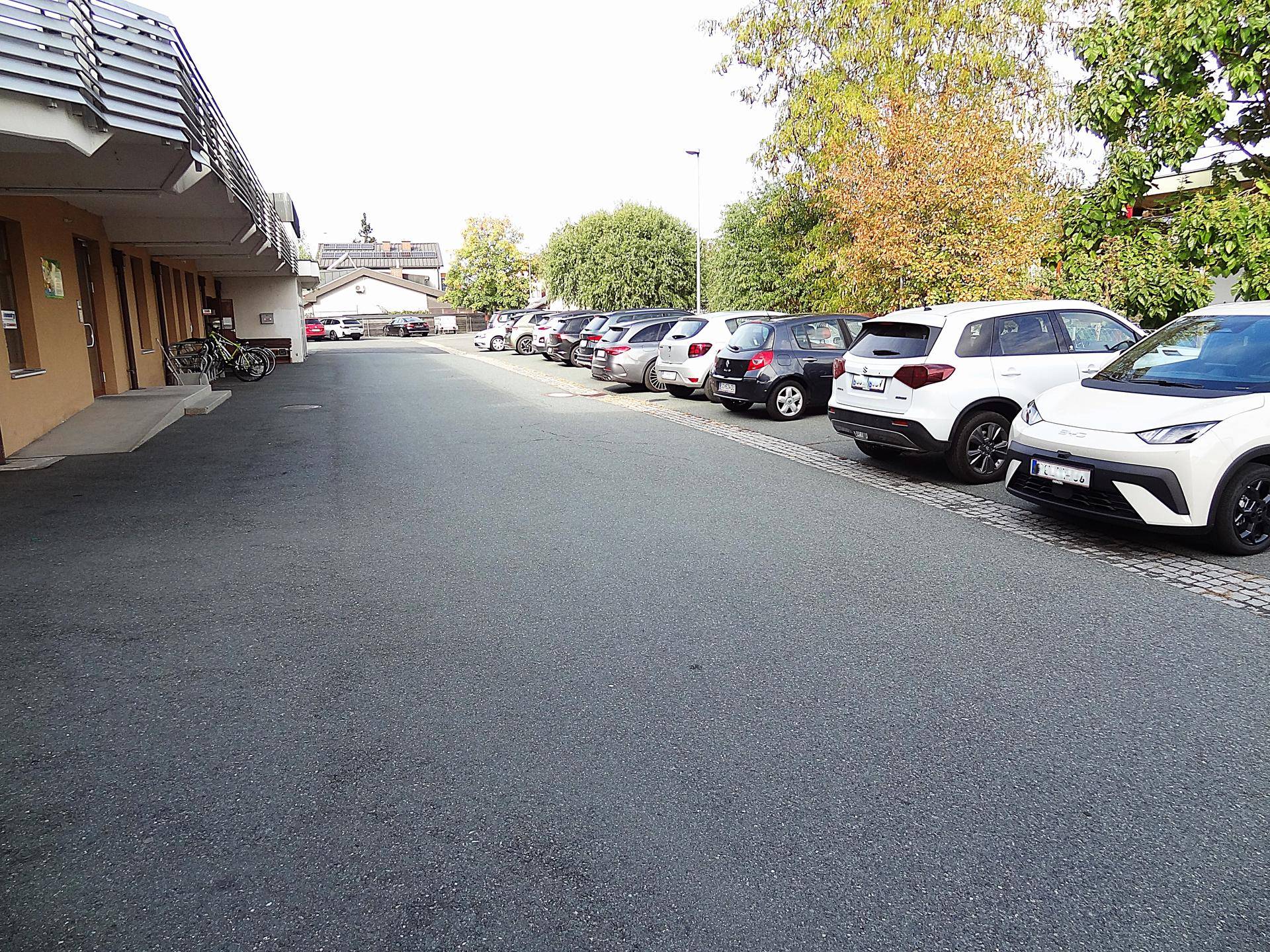 Langer asphaltierter Parkplatz mit geparkten Autos entlang der Gebäudefassade.