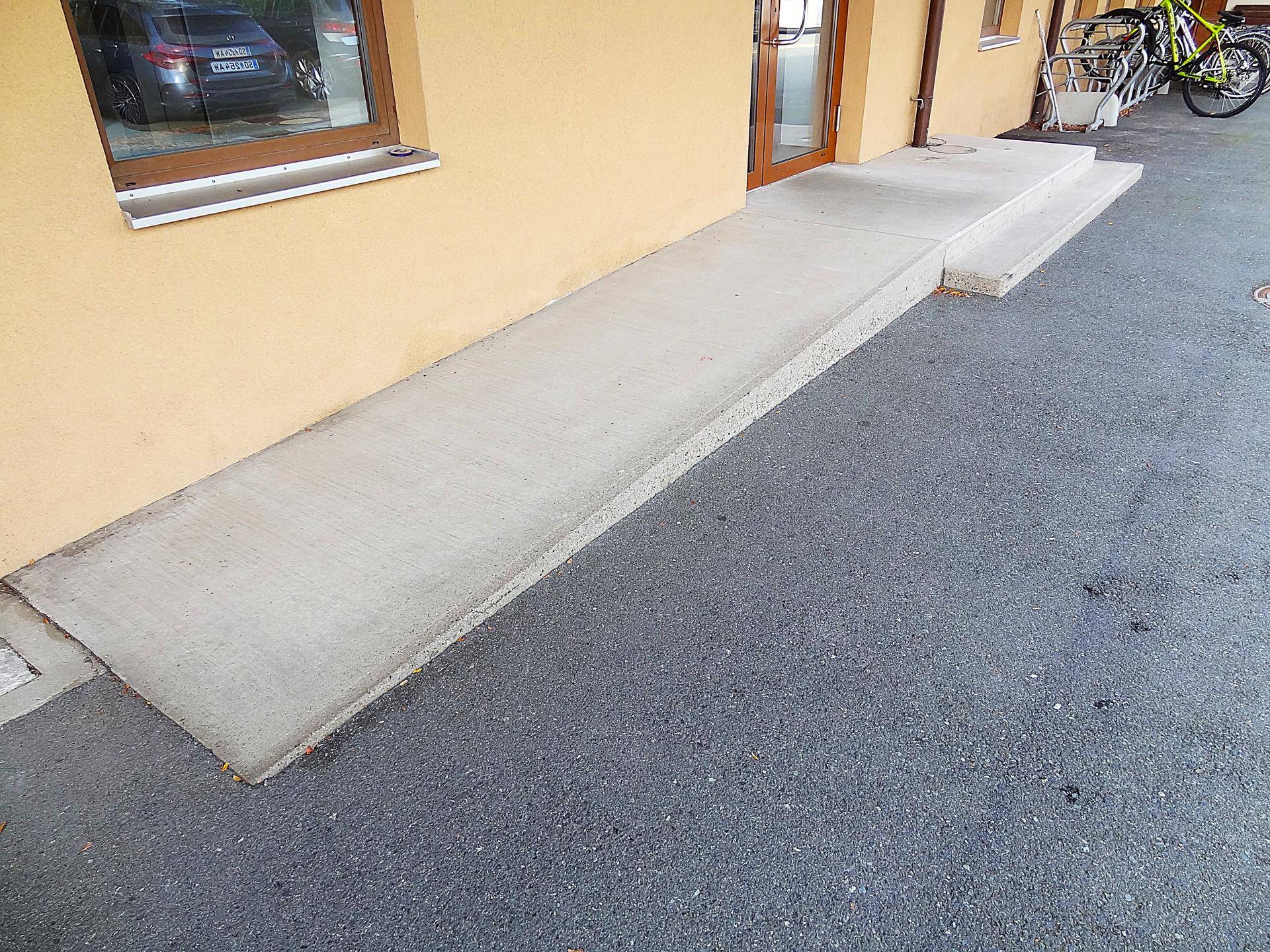 Lange Betonrampe und Stufen führen zum Gebäudeeingang, mit Fahrradabstellplatz im Hintergrund.