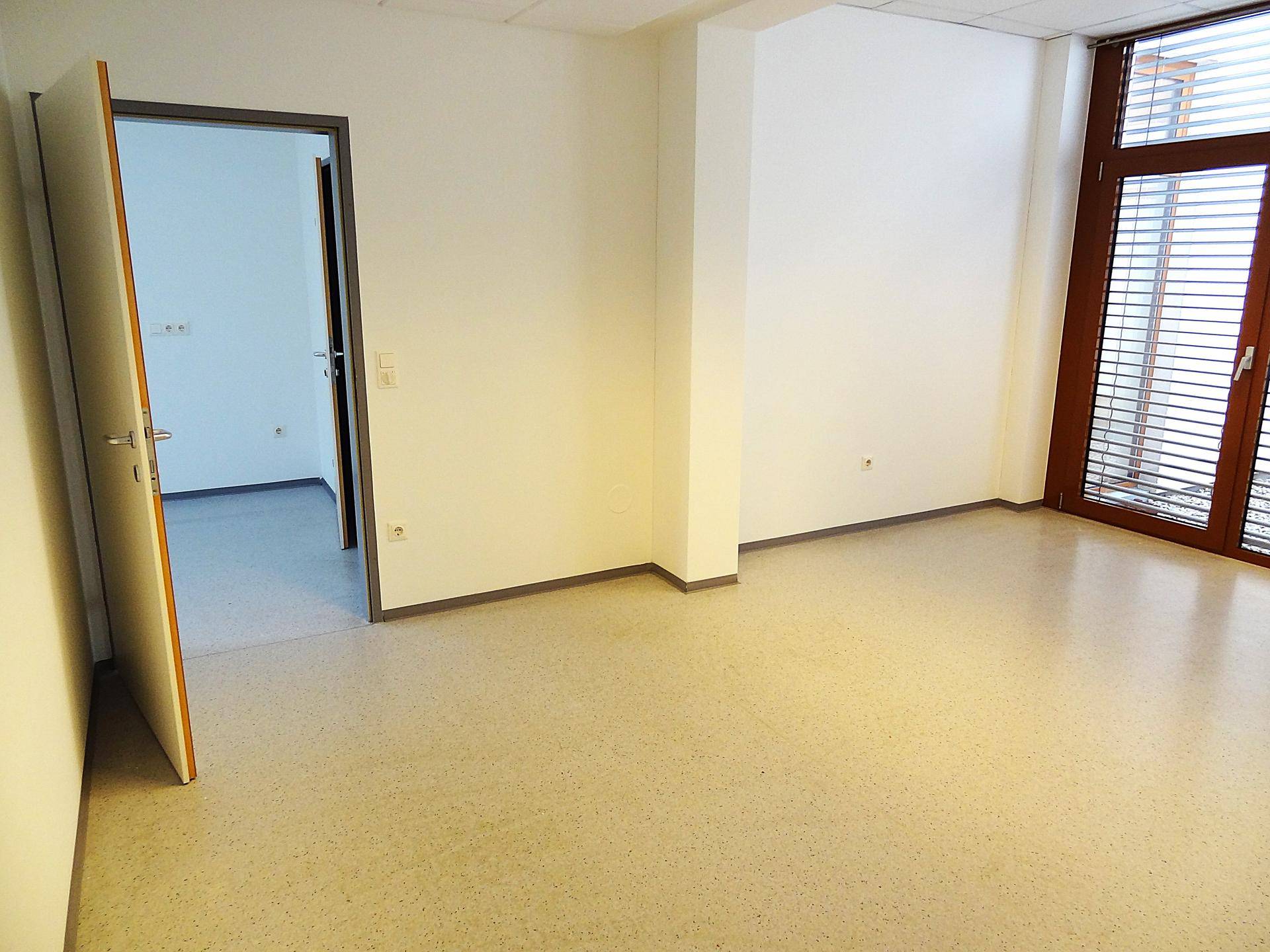 Heller Büroraum mit hellem Boden, großen Fenstern mit Jalousien und offener Tür.