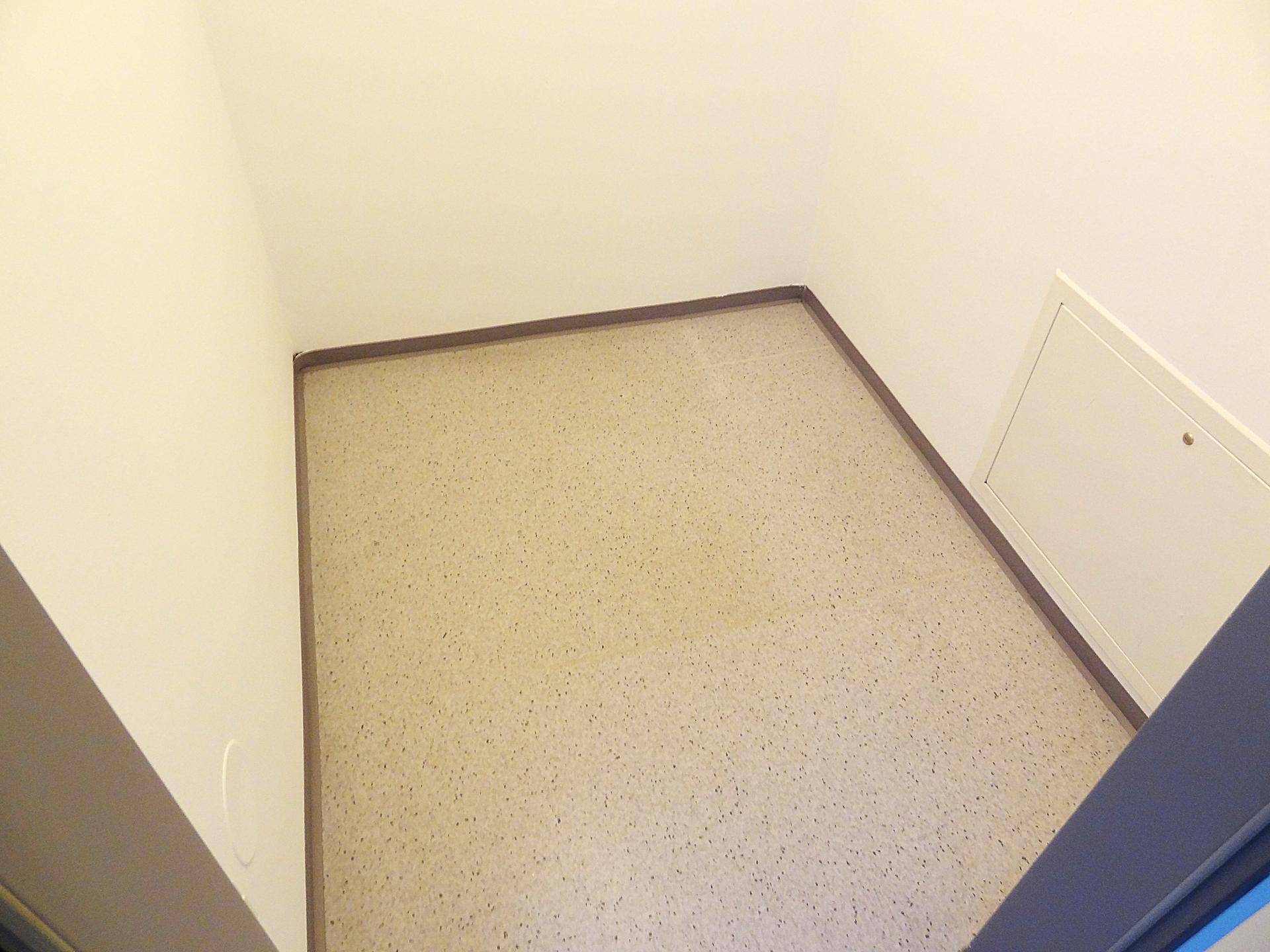 Kleiner, leerer Raum mit hellem Boden und einer Wartungsklappe an der Wand.