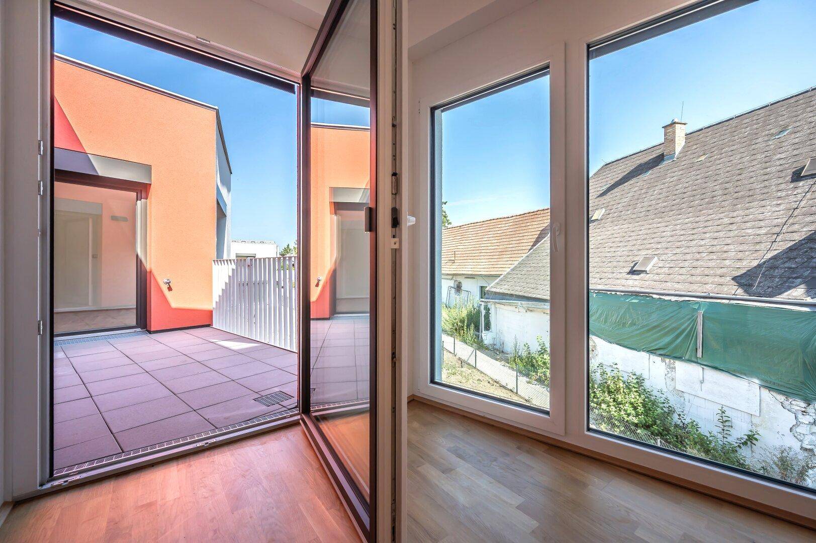 Heller Innenraum mit großen Fenstern und Blick auf die geflieste Terrasse und die umliegenden Dächer.