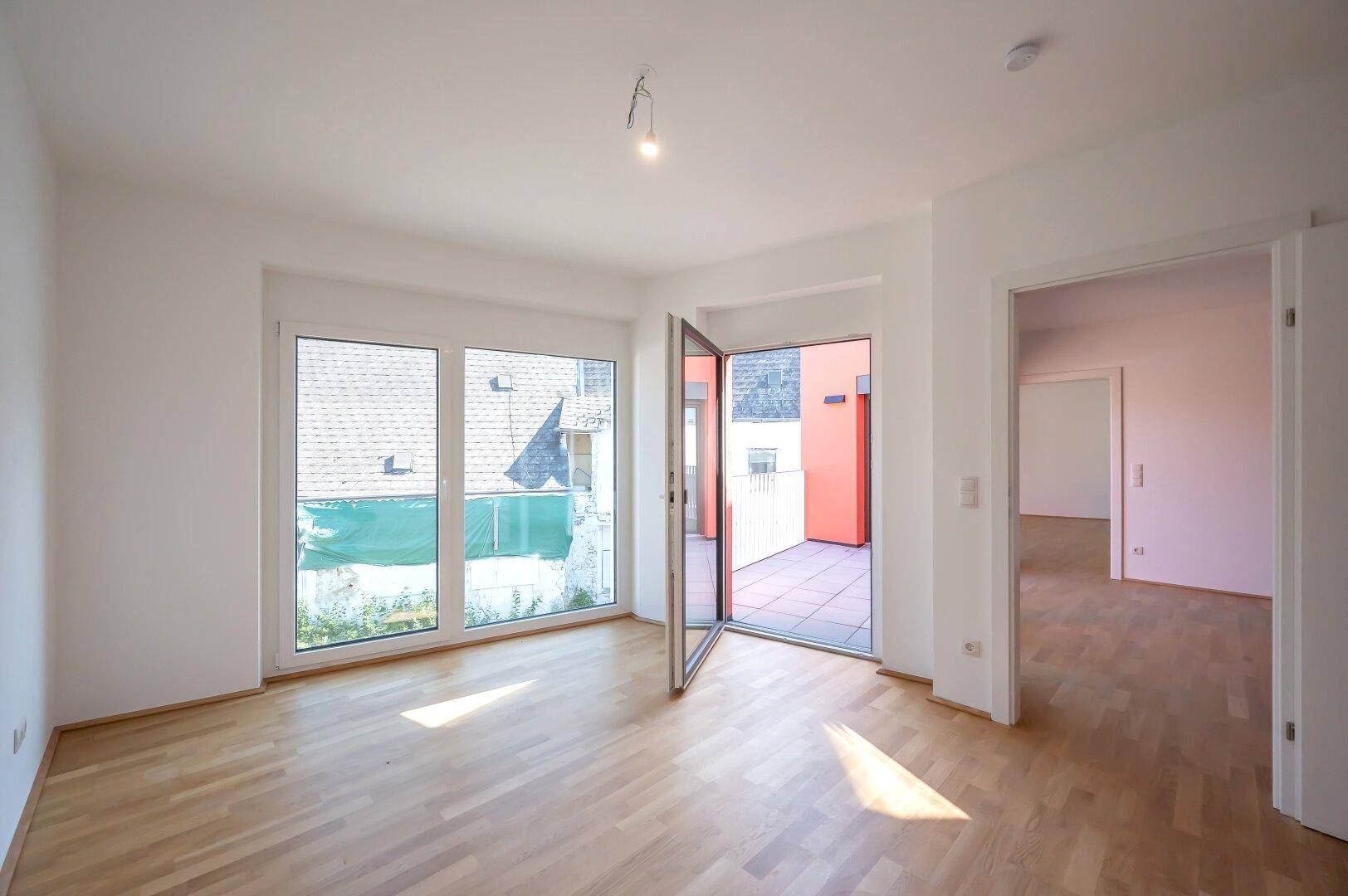 Lichtdurchfluteter Raum mit Parkettboden und großen Fenstern, die den Blick nach draußen freigeben.