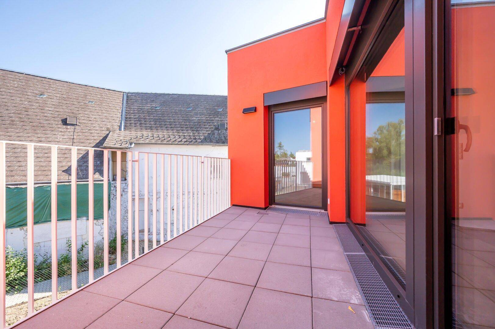 Sonniger Terrassenbereich mit Fliesen und modernem Geländer, angrenzend an ein rotes Gebäude.