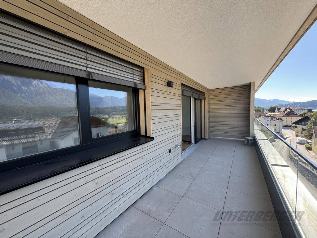 Langer, überdachter Balkon mit heller Holzfassade, grauen Bodenfliesen und Glasgeländer
