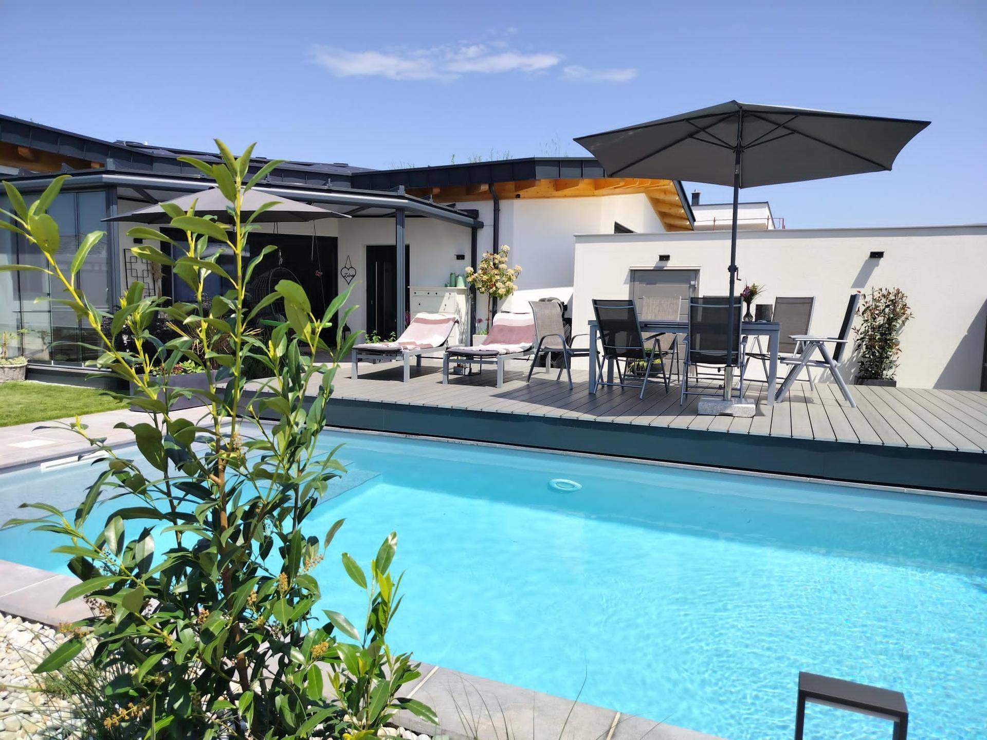 Sonniger Außenbereich mit Pool, Holzterrasse und Gartenmöbeln, umgeben von Grün.