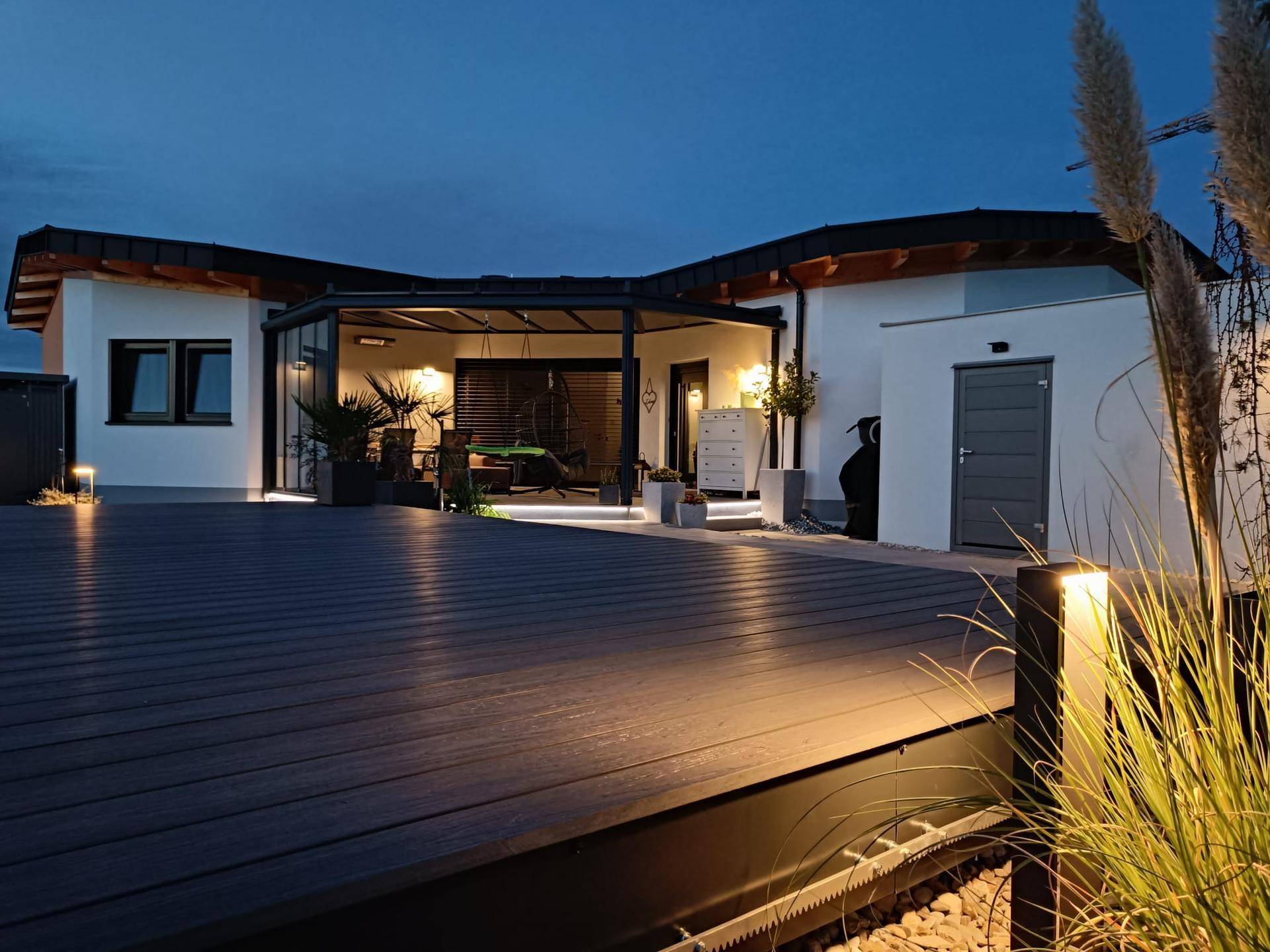 Beleuchtete Holzterrasse am Abend mit angrenzendem Wintergarten und moderner Architektur.