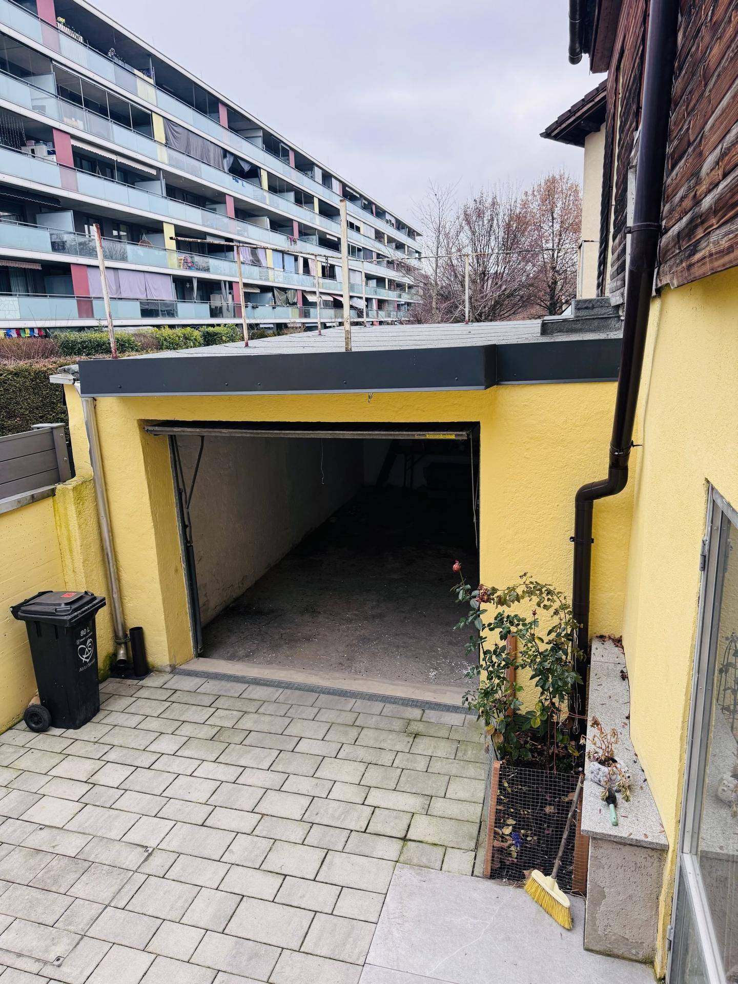 Offene Garage mit gepflastertem Vorplatz und einem Mehrfamilienhaus im Hintergrund.