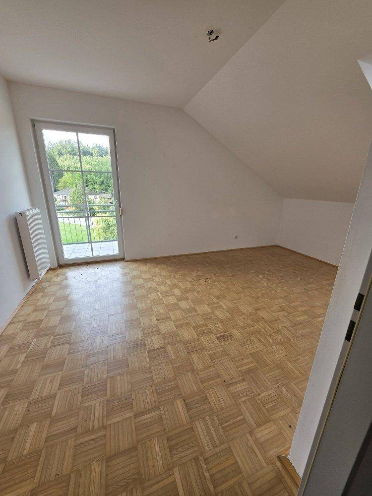 Helles Zimmer mit Parkettboden, Dachschräge und Zugang zu einem kleinen Balkon.