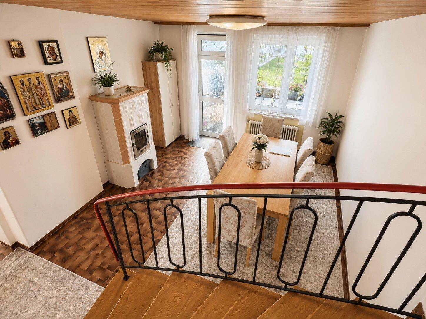 Gemütlicher Essbereich mit Holztisch, Kachelofen und Blick auf den Garten durch große Fenster.