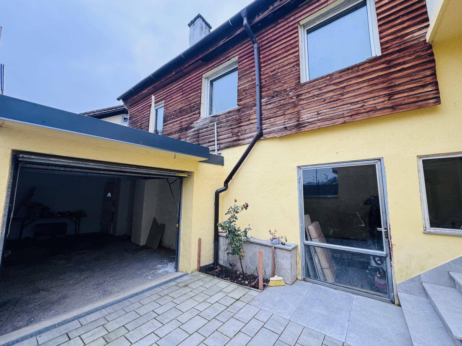 Seitenansicht des Hauses mit Garage, gepflastertem Bereich und Holzverkleidung im Obergeschoss.