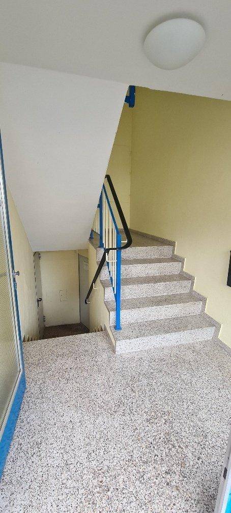 Treppenhaus mit Steinfliesenboden, gelben Wänden und blauem Geländer in einem Mehrfamilienhaus.