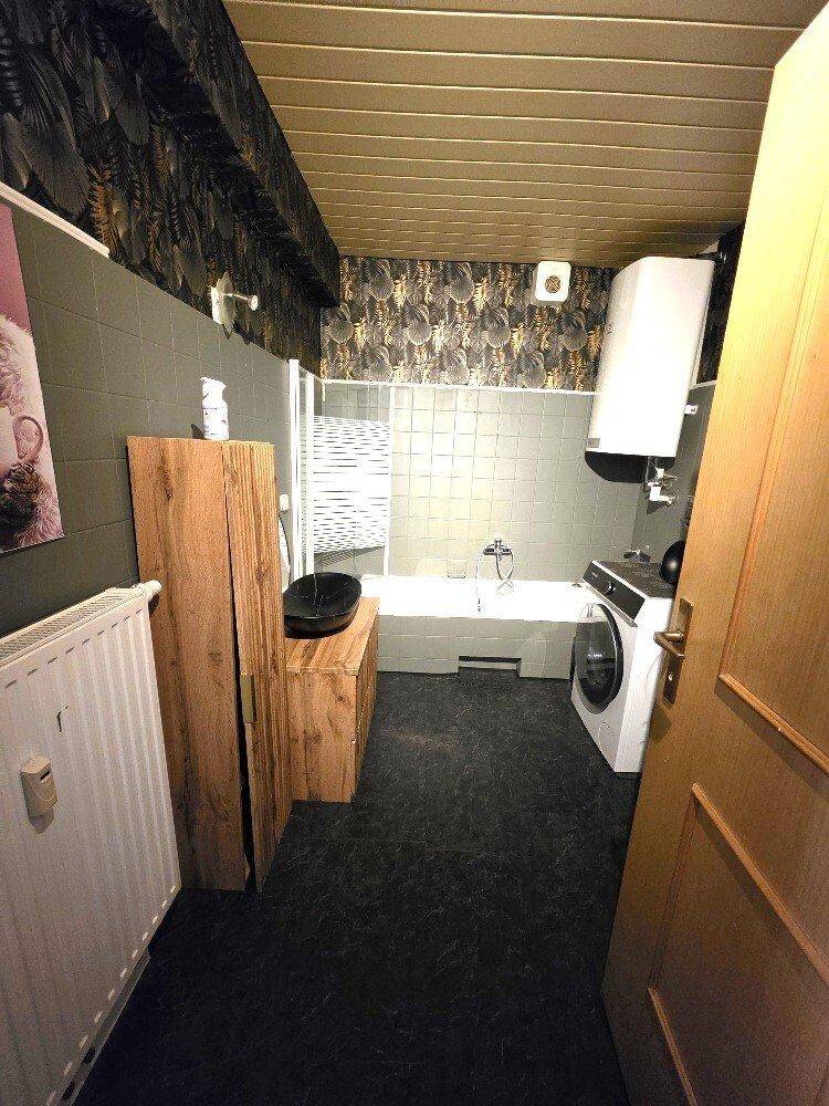 Badezimmer mit Badewanne, Waschmaschine und dunklen Fliesen, ergänzt durch Holzmöbel.