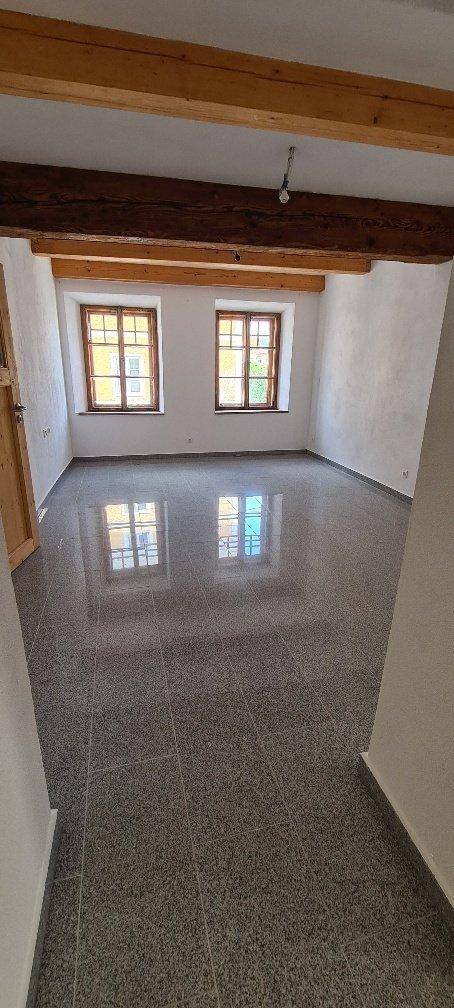 Heller Wohnraum mit Holzbalken, zwei Fenstern und Fliesenboden, der viel Licht reflektiert.