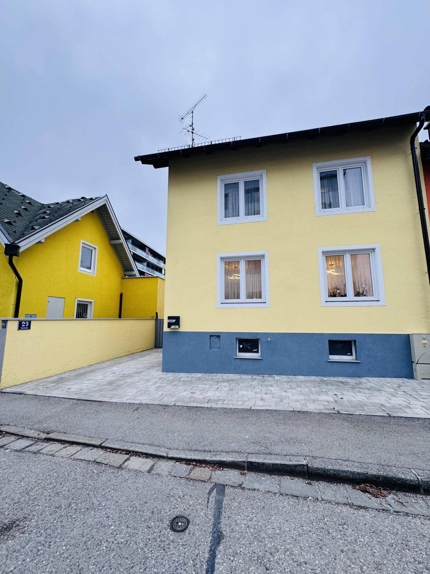 Vorderansicht des gelben Hauses von der Straße, mit gepflasterter Einfahrt und Nachbargebäude.