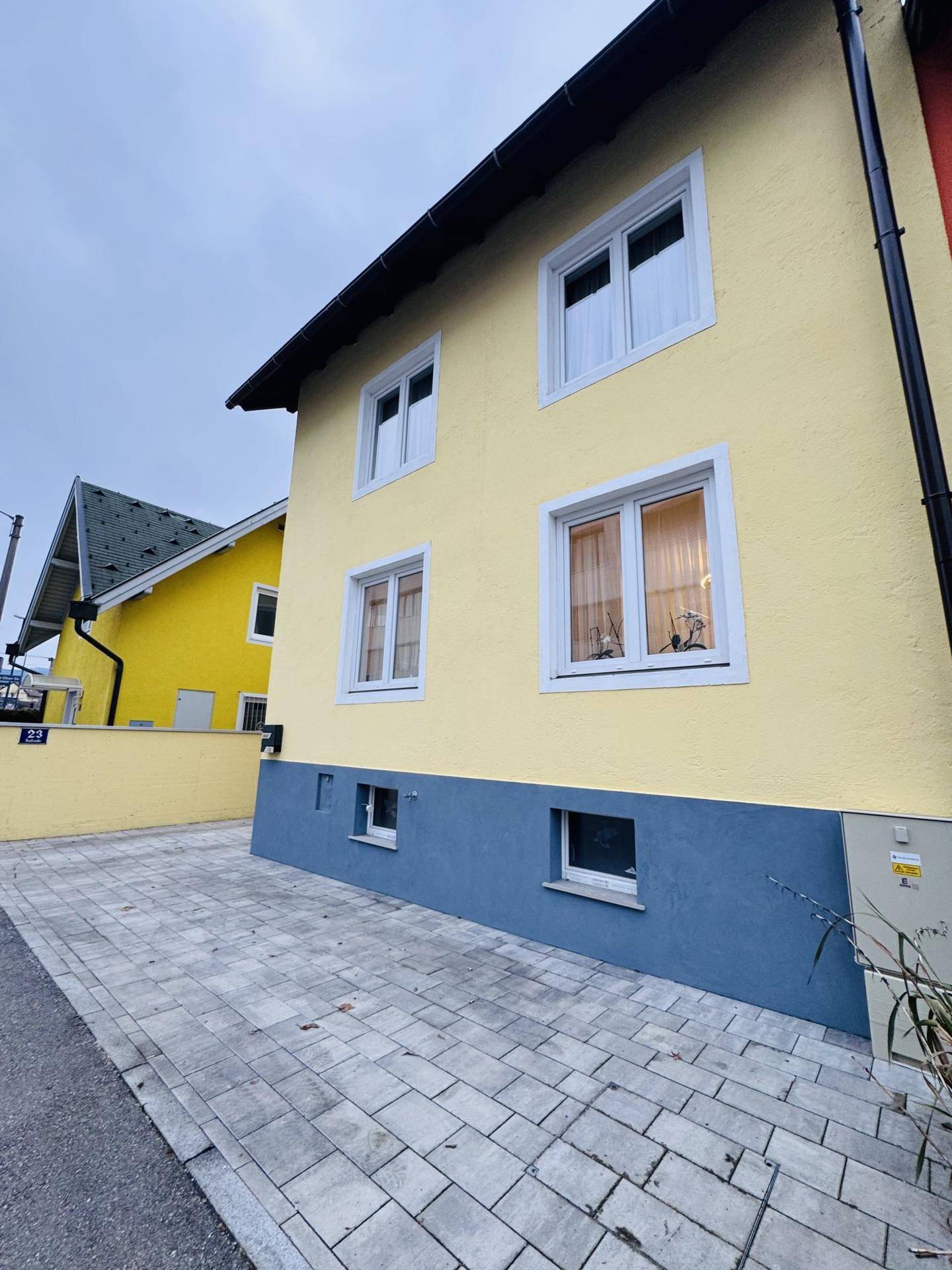 Seitenansicht des gelben Hauses mit gepflasterter Einfahrt und einem weiteren gelben Gebäude im Hintergrund.