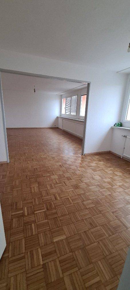 Geräumiges, leeres Wohnzimmer mit Fischgrätparkett und offenem Durchgang zu einem weiteren Raum.